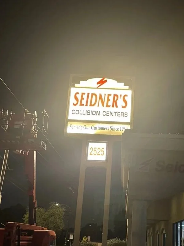 A tall illuminated sign for Seidner's Collision Centers at night, displaying the address 2525, with a lightning bolt symbol and tagline 'Serving Our Customers Since 1964'.