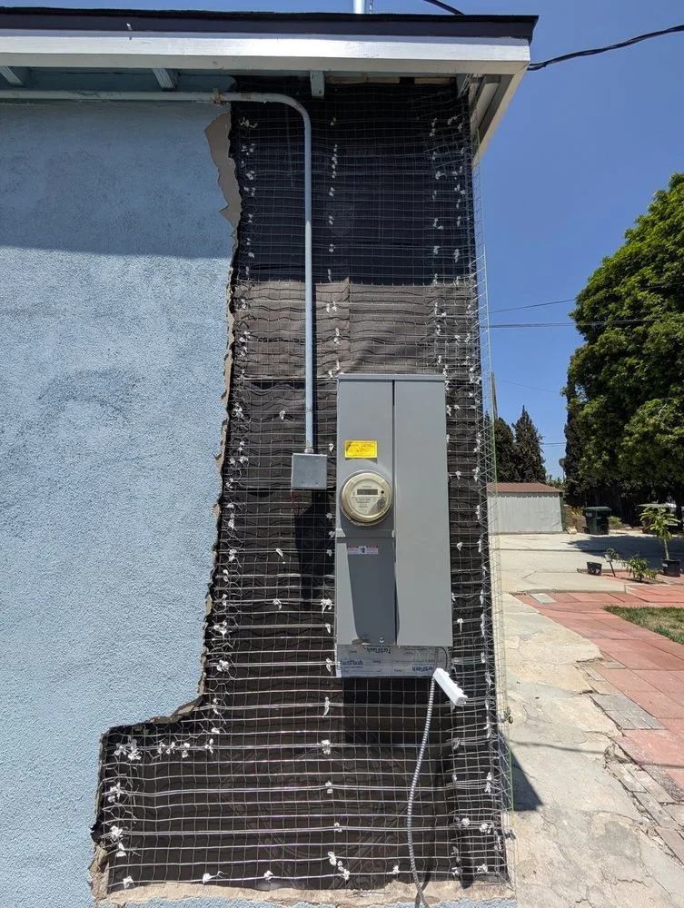 Exterior wall of a building under renovation, with part of the wall removed, exposing wiring and an electrical meter box. Wrapping mesh surrounds the exposed area. A sidewalk and trees are visible in the background.