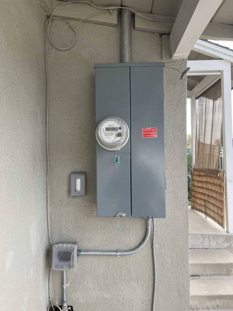 An electrical meter box mounted on a beige stucco wall next to an outdoor porch area. The meter has a round display and a small green key hanging from it. Below it, a conduit runs into a small frost-proof electrical outlet box, and a power outlet is mounted on the wall to the left.