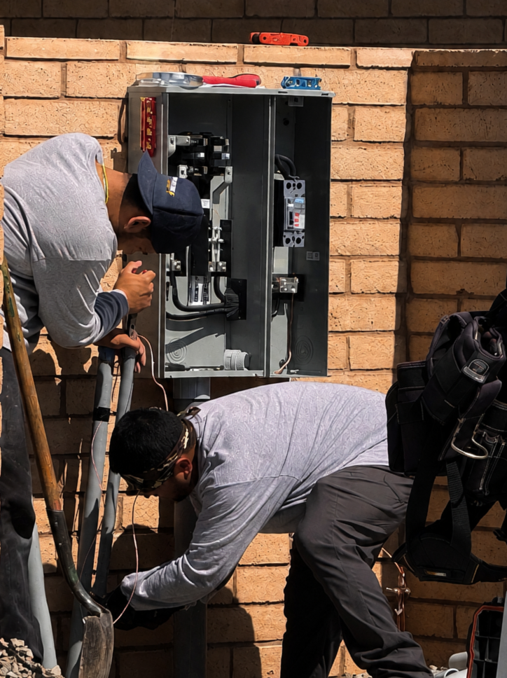 Two electricians installing or repairing an electrical panel on a brick wall outside.