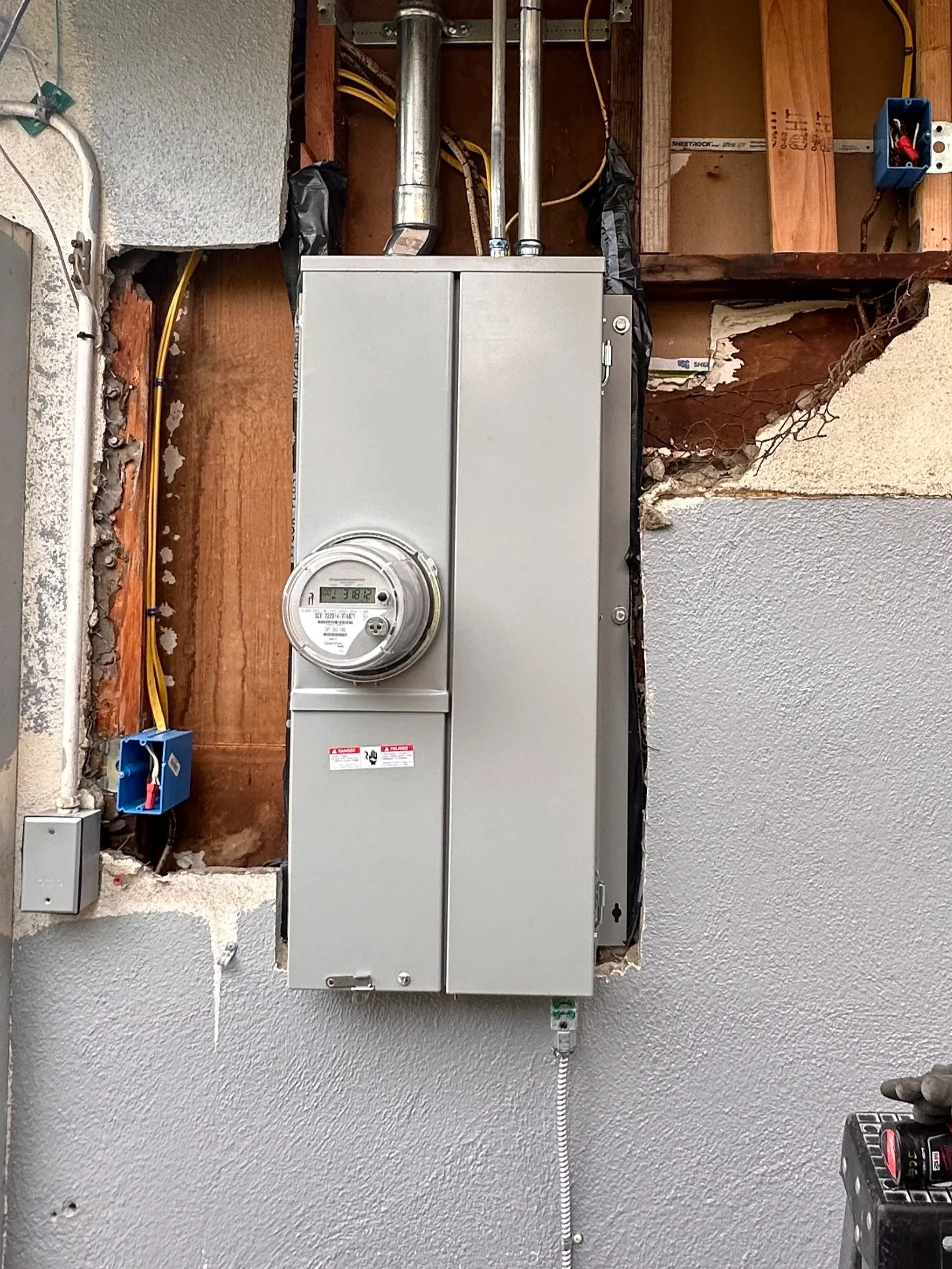 An electrical meter mounted on exterior wall next to an unfinished section of wall with exposed wooden studs and electrical wiring, which is partially covered with drywall and textured paint.