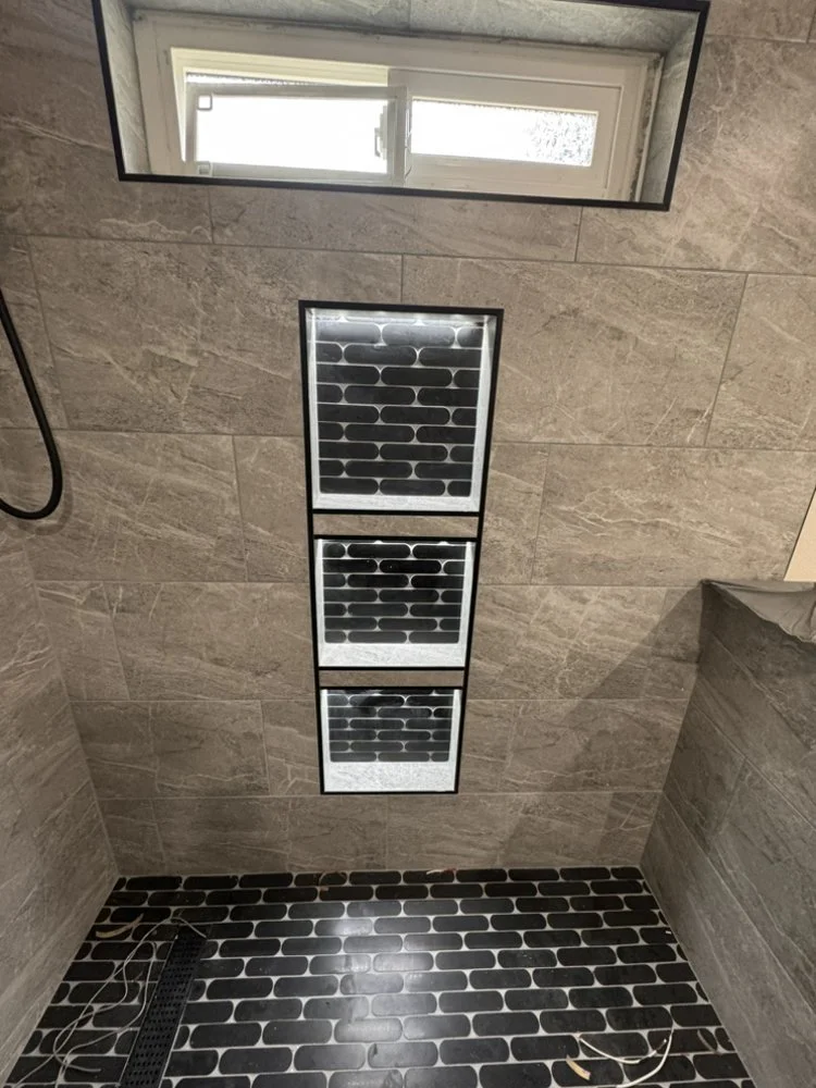 Bathroom ceiling with three rectangular air vents or lights, a window above, and black and gray tile flooring and wall tiles.