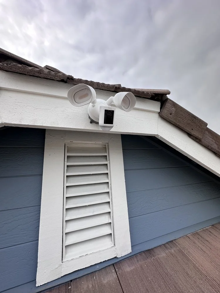 A security camera with wind turbine-like attachments mounted on the eaves of a house with blue siding and a white vent, under a cloudy sky.