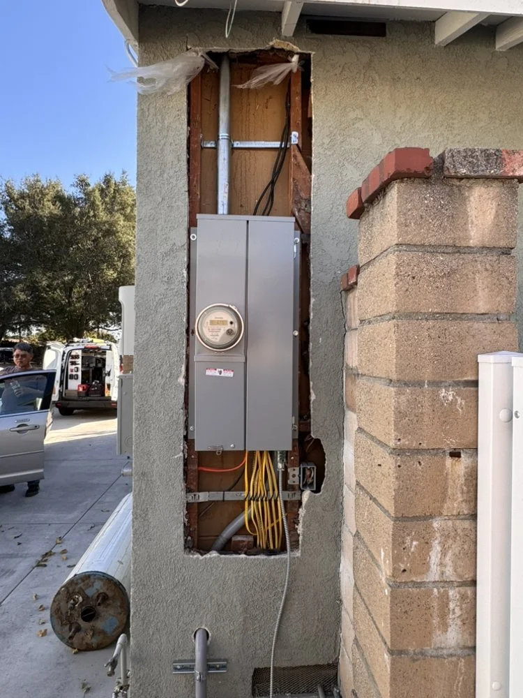 Partial view of electrical wiring and meter box on exterior wall of building, with a brick wall on the right and a sidewalk with a person and vehicle in the background.