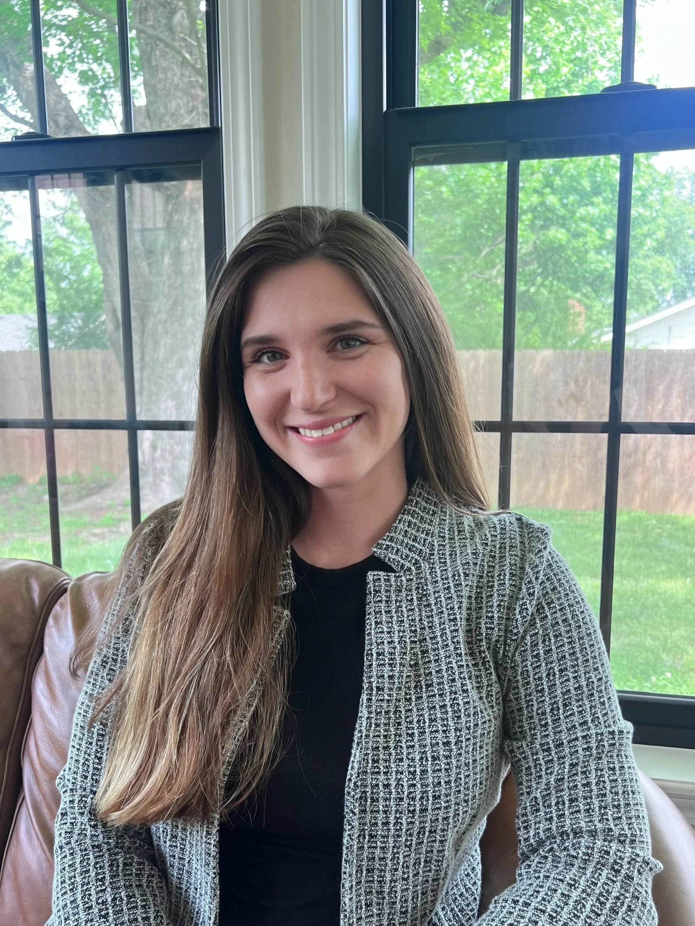 A young woman with long brown hair, smiling, sitting indoors in front of large windows with greenery outside.