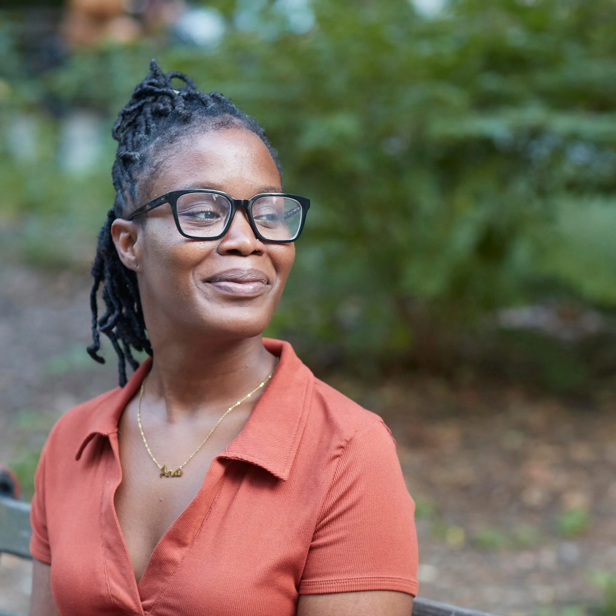 A woman with glasses and braided hair smiling outdoors with greenery in the background, wearing an orange collared shirt and a gold necklace.