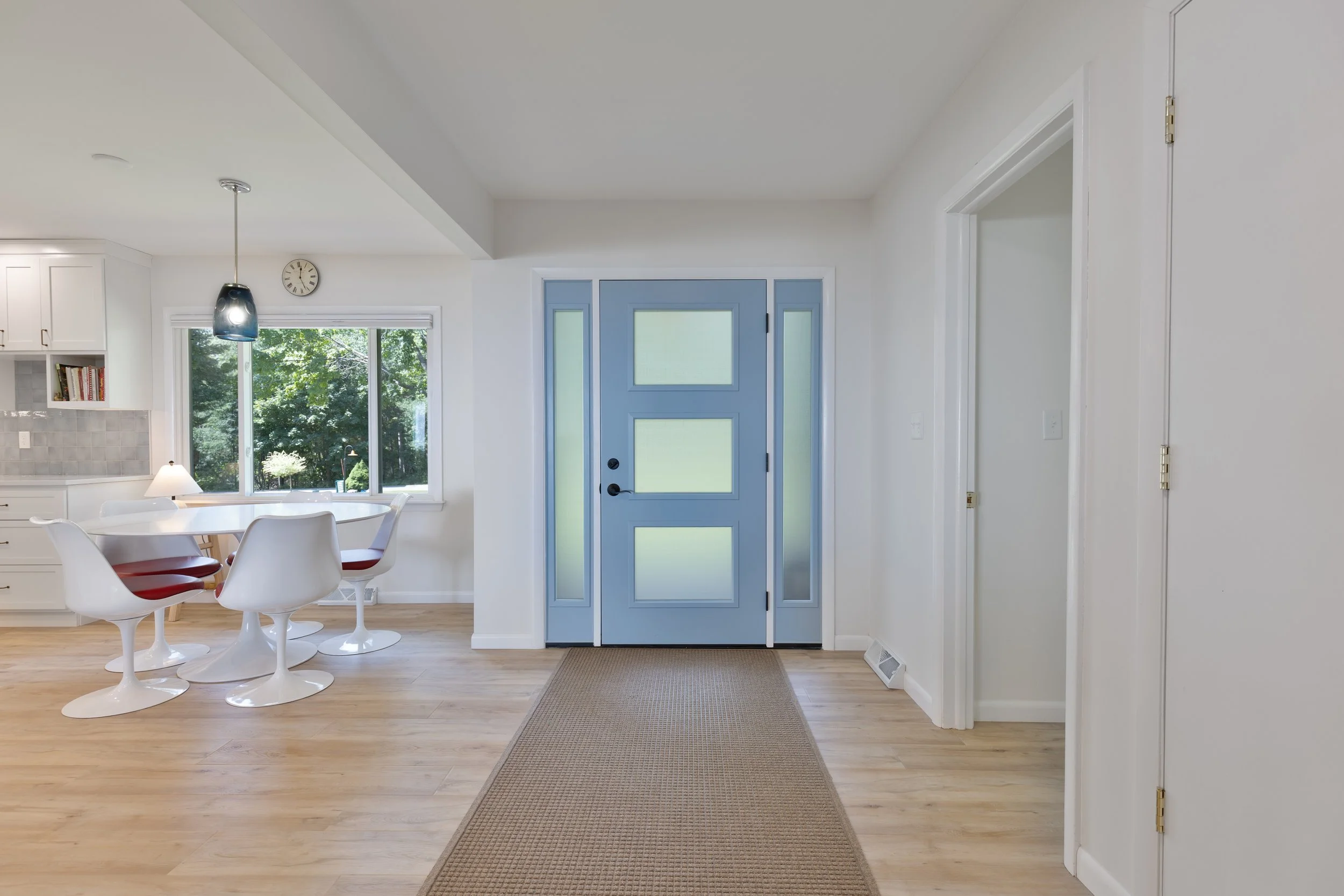 Interior view of a modern house entrance with a blue front door, a kitchen visible on the left, a dining area with white chairs, a window with greenery outside, and a hallway on the right.