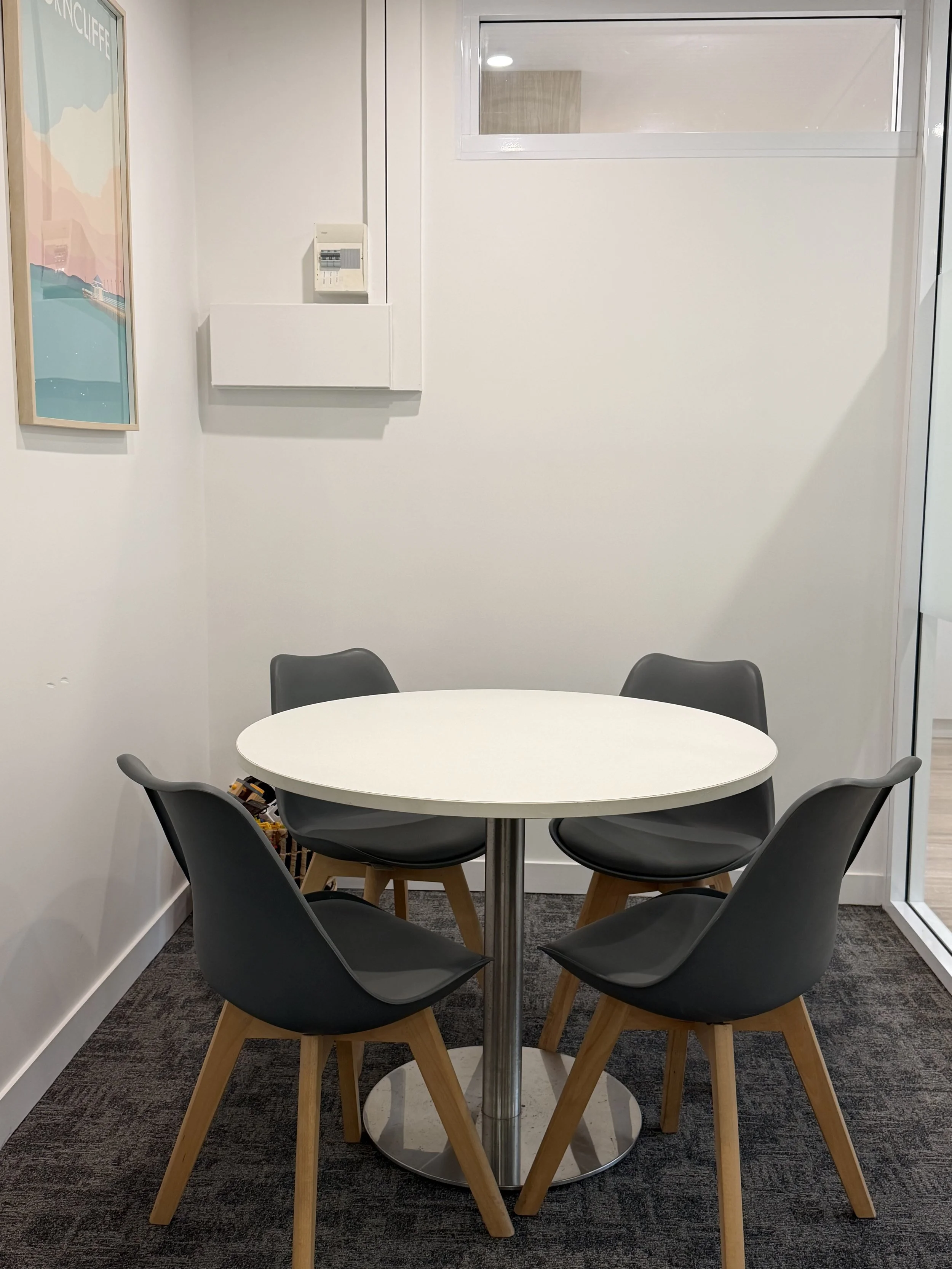 A round white table with six black chairs in a small conference room or office break area. There is a framed colorful map or artwork on the left wall and a small window on the upper right wall. The floor is dark carpeted.