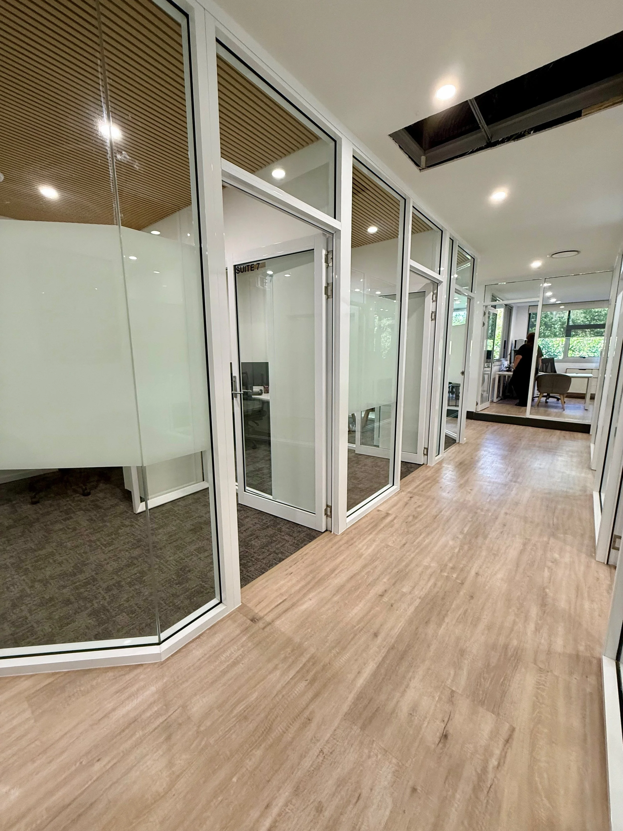 Modern office space with glass partitions, wooden flooring, and a woman working at a desk near large windows.