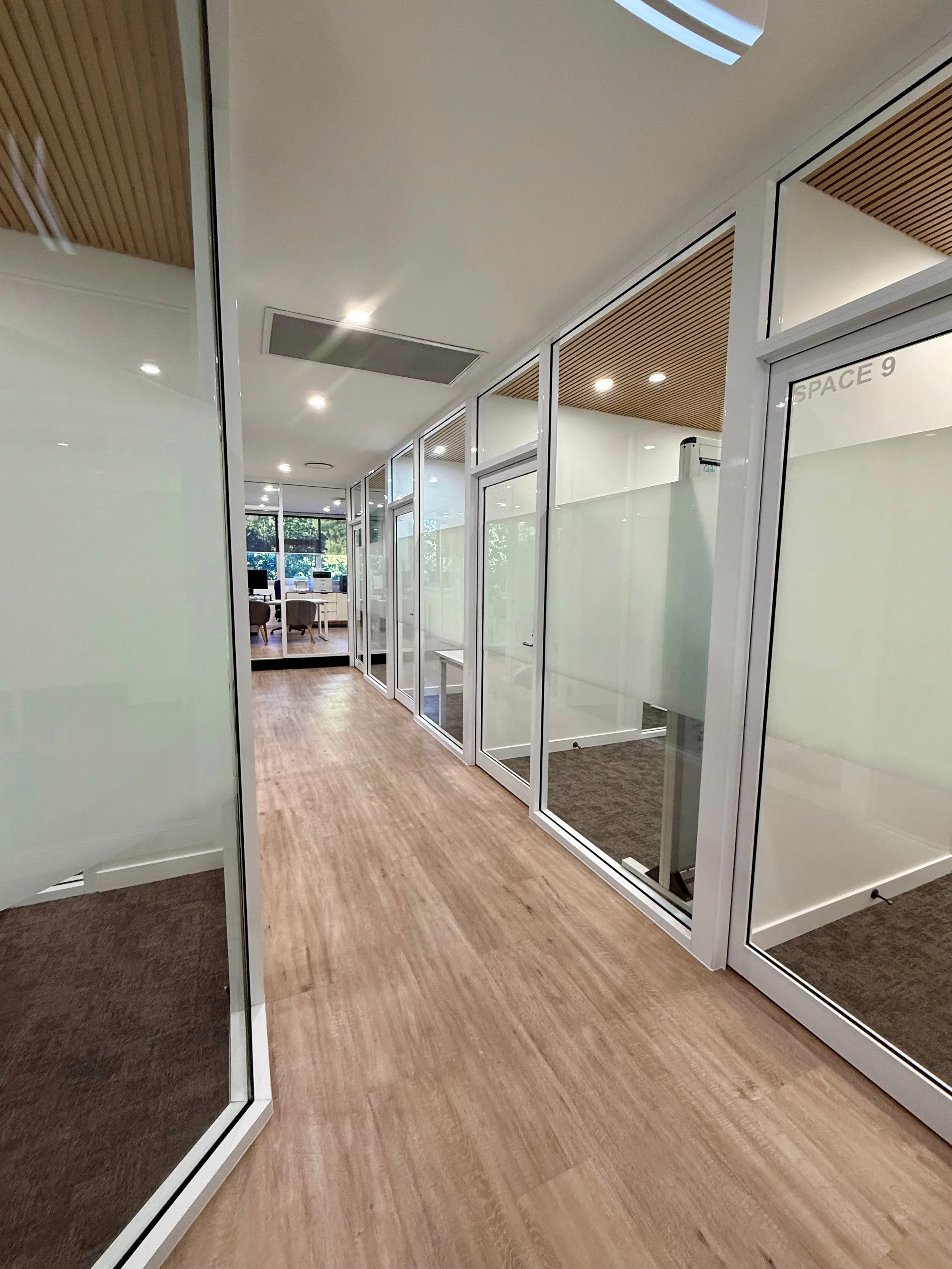Modern office corridor with glass partitions, wooden flooring, and ceiling with wooden slats, leading to a room with chairs, desks, and computer monitors.