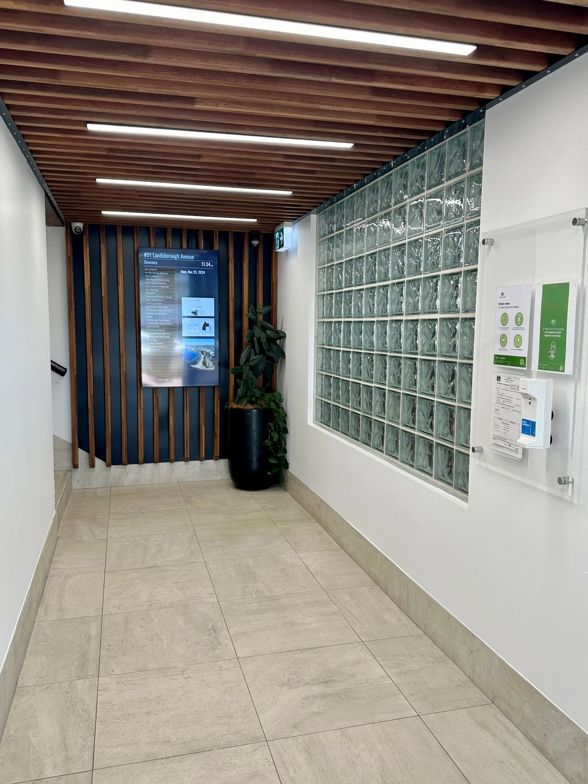 Interior corridor of a building with a wooden slatted ceiling, glass block wall, digital directory, potted plant, white walls, and beige tiled floor.