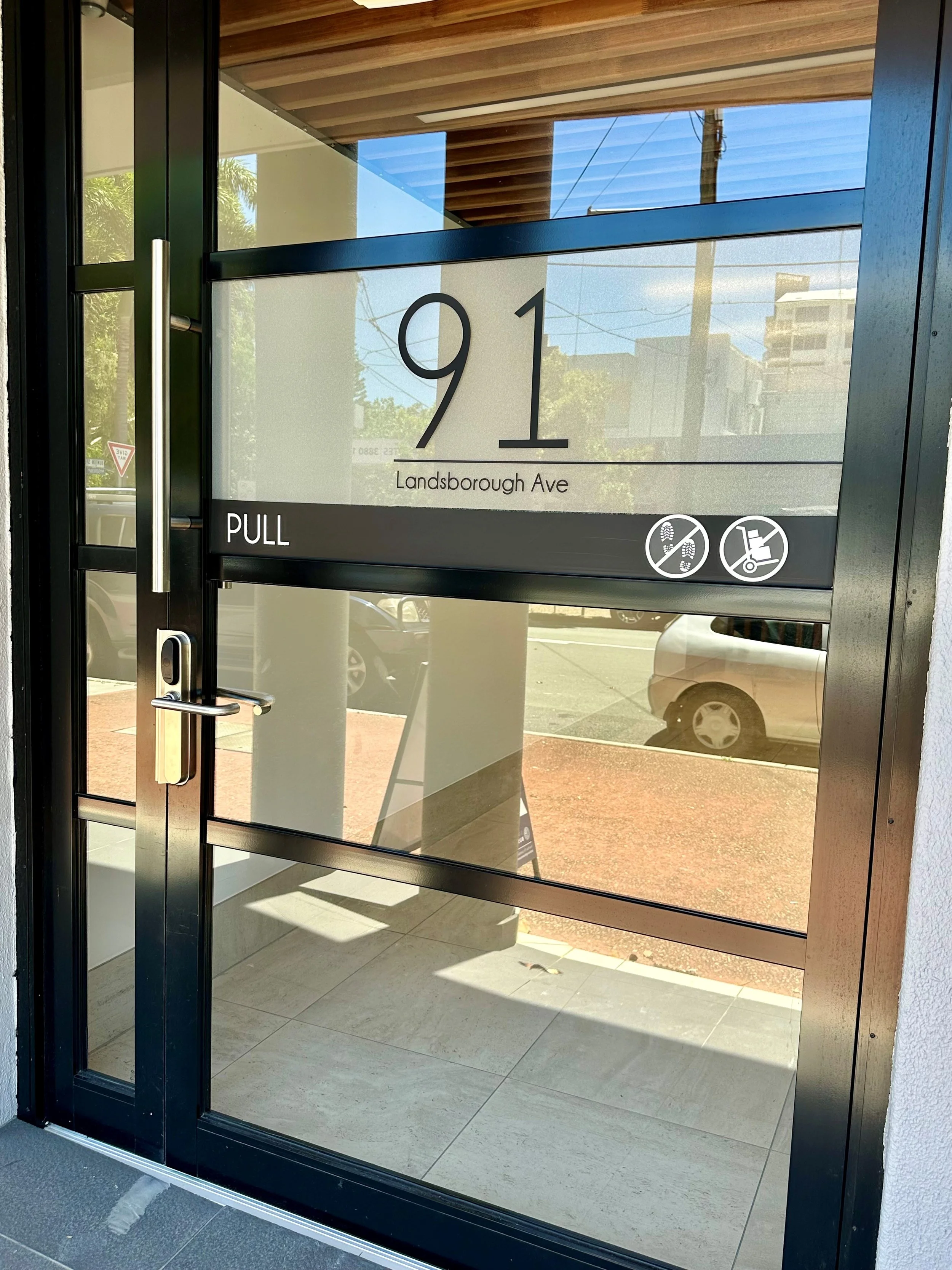 Glass door with black frame displaying address 91 Landsborough Ave, with a silver handle, and close-up of the door frame, showing reflections of street and parked cars outside.