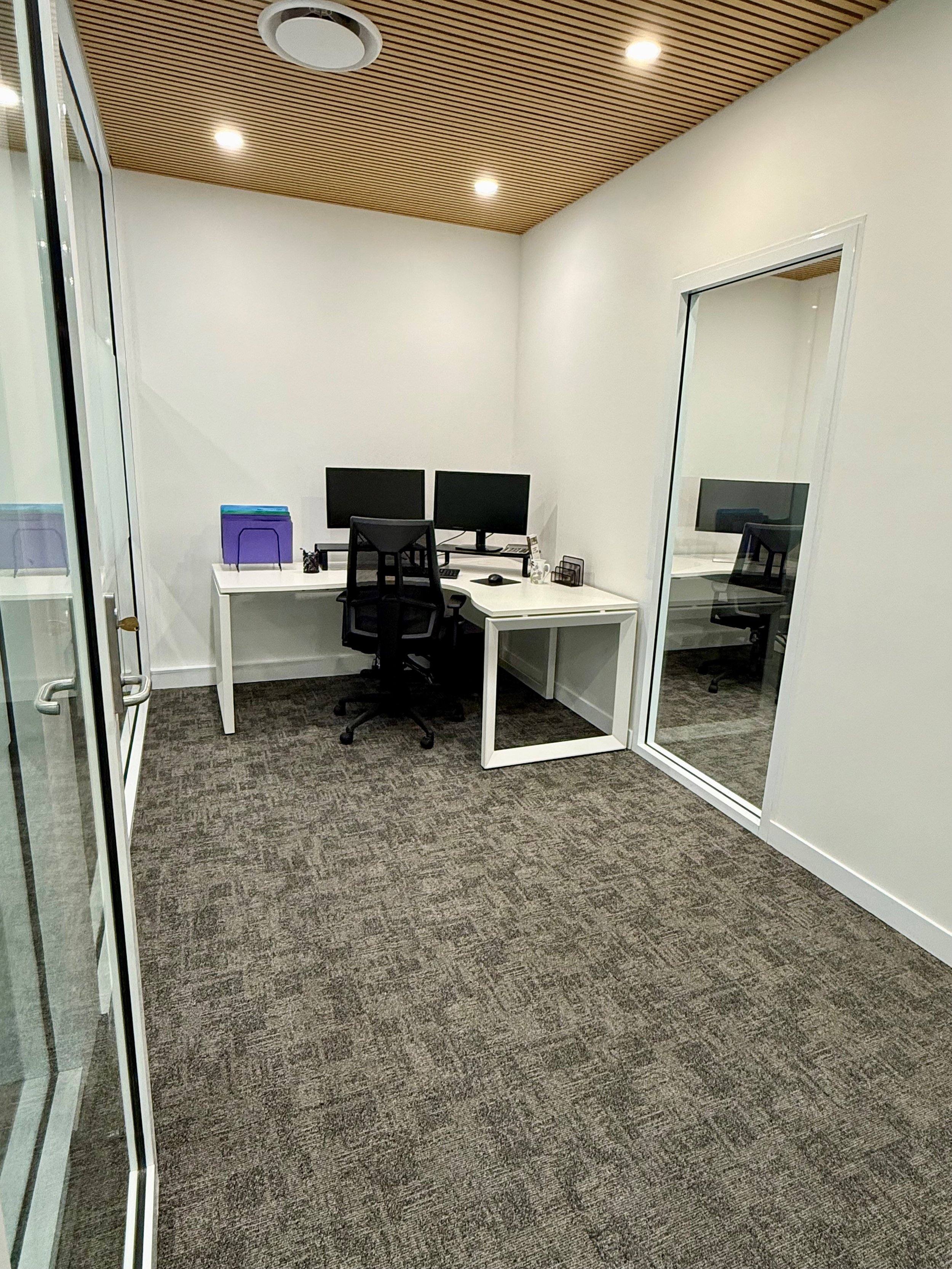 An office room with a white desk, two monitors, a black office chair, a purple file folder, and a glass door with a window. The room has a brown carpeted floor, white walls, and a wooden slatted ceiling with four recessed lights.