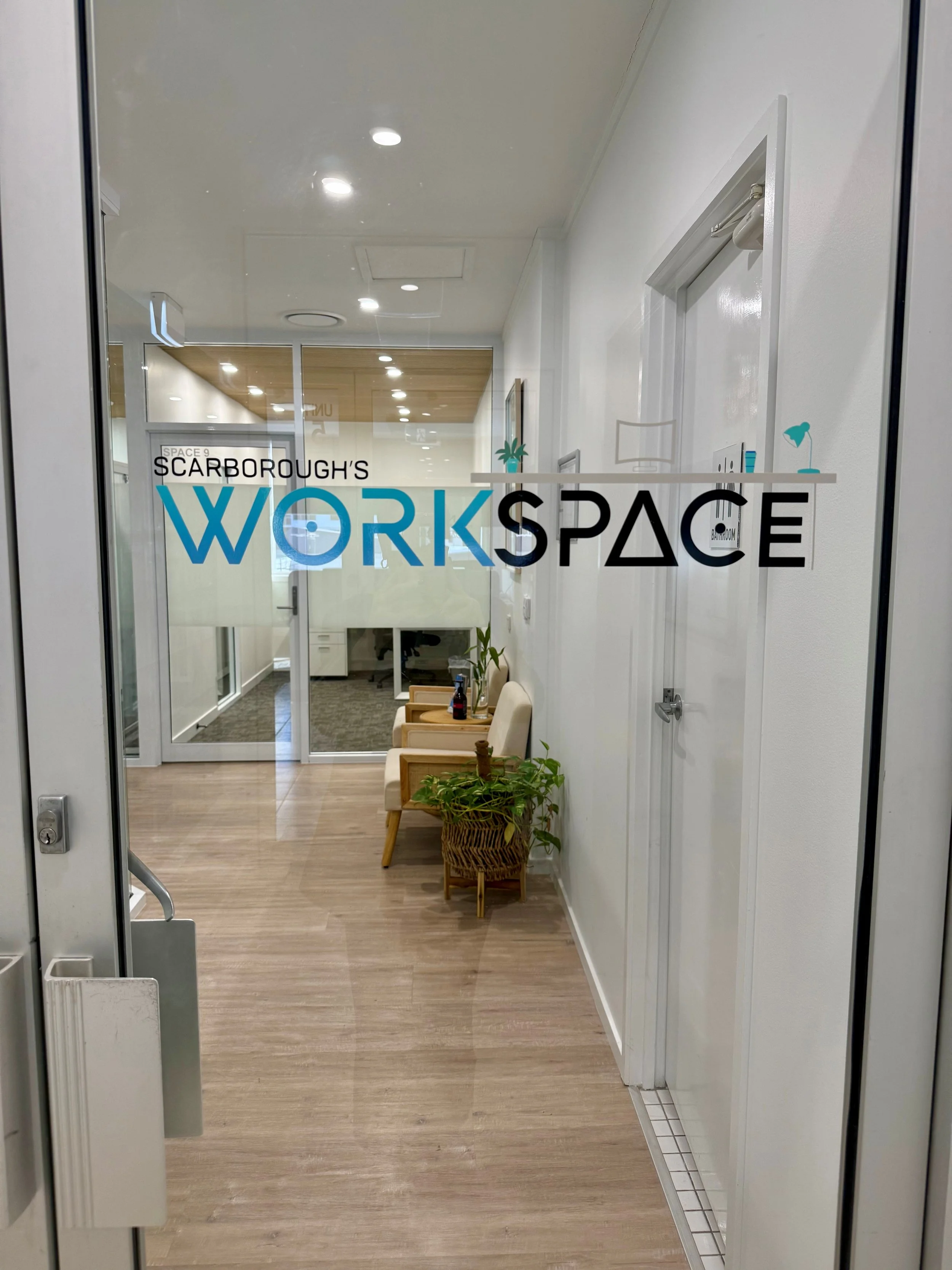 View through glass door into a modern, well-lit office workspace with wooden flooring, a seating area with chairs and a table, plants, and white buildings and glass walls in the background.