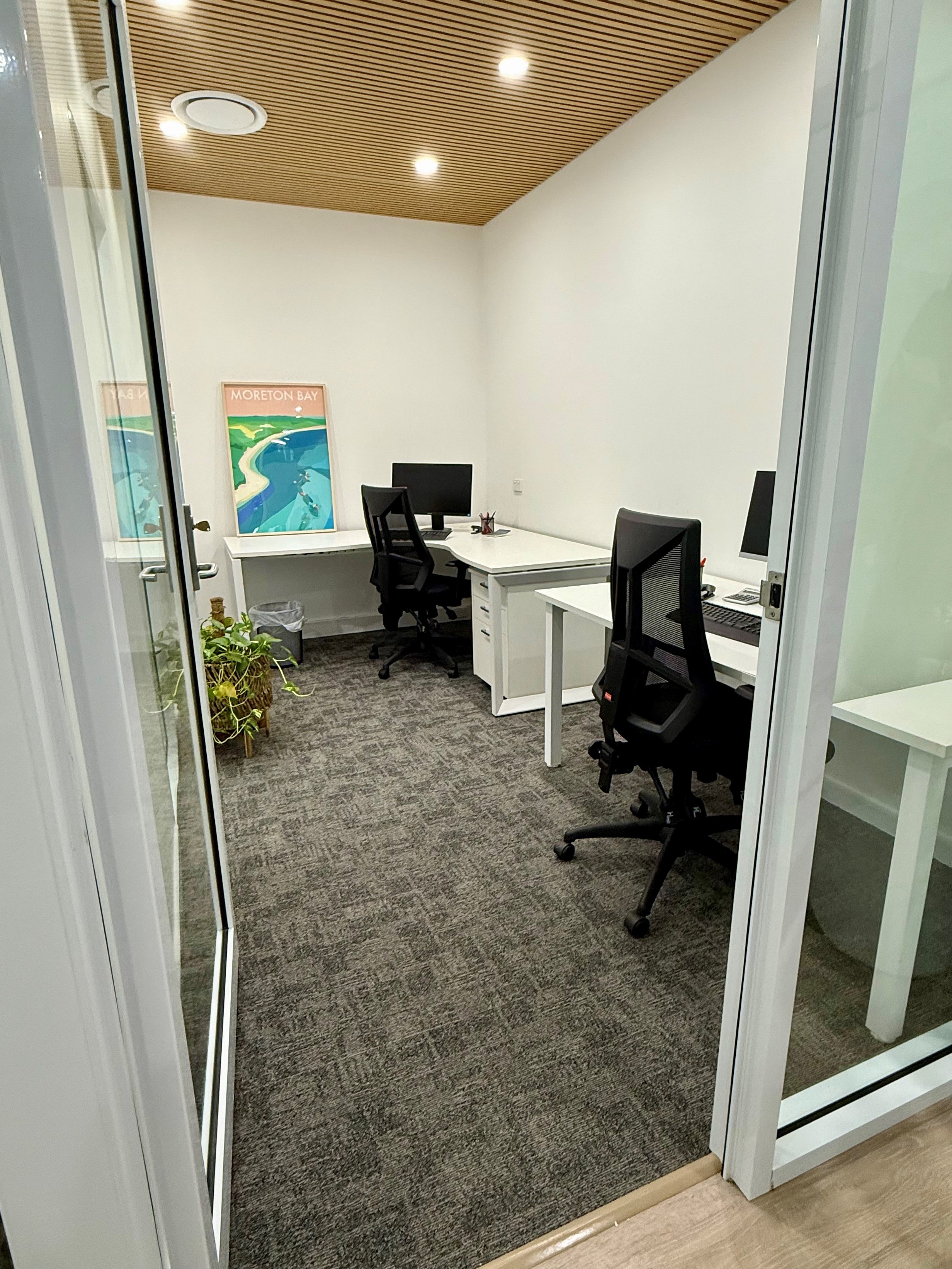 An office space with two black office chairs, white desks, a framed colorful artwork on the wall, a green plant in a basket, and a window with glass door leading to the room. The ceiling has wooden panels with recessed lighting.