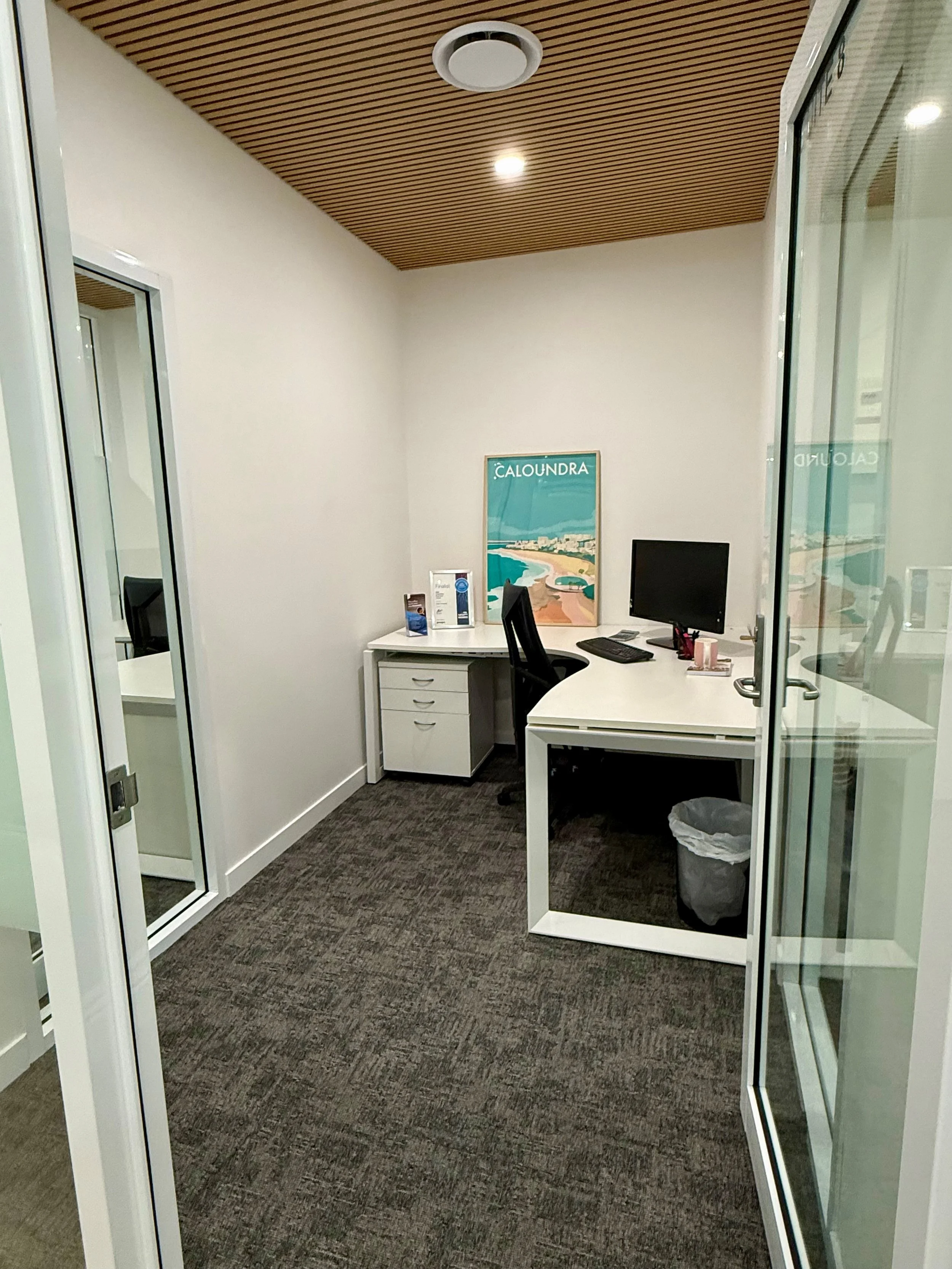 An empty office space with white walls, a wooden ceiling, a small white desk with a black swivel chair, a computer monitor, and a trash bin. There's a framed poster of a beach scene and some brochures on the desk. The room is viewed through a glass d