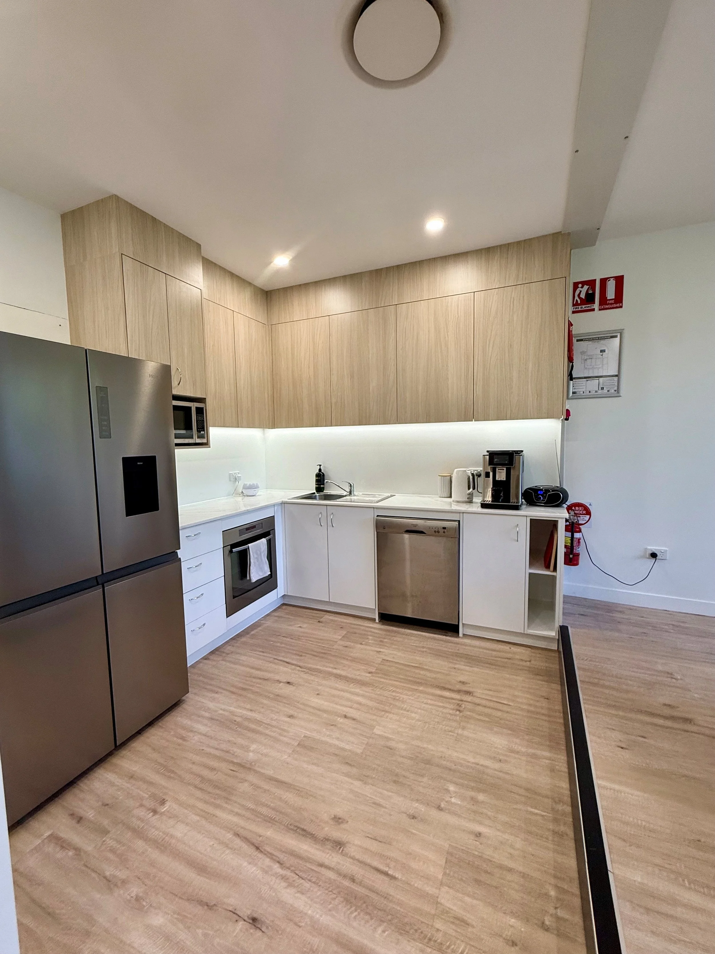 Modern kitchen with white cabinets, wood upper cabinets, stainless steel refrigerator, stove, microwave, small appliance on counter, built-in sink, fire safety equipment on wall, and wood-look flooring.