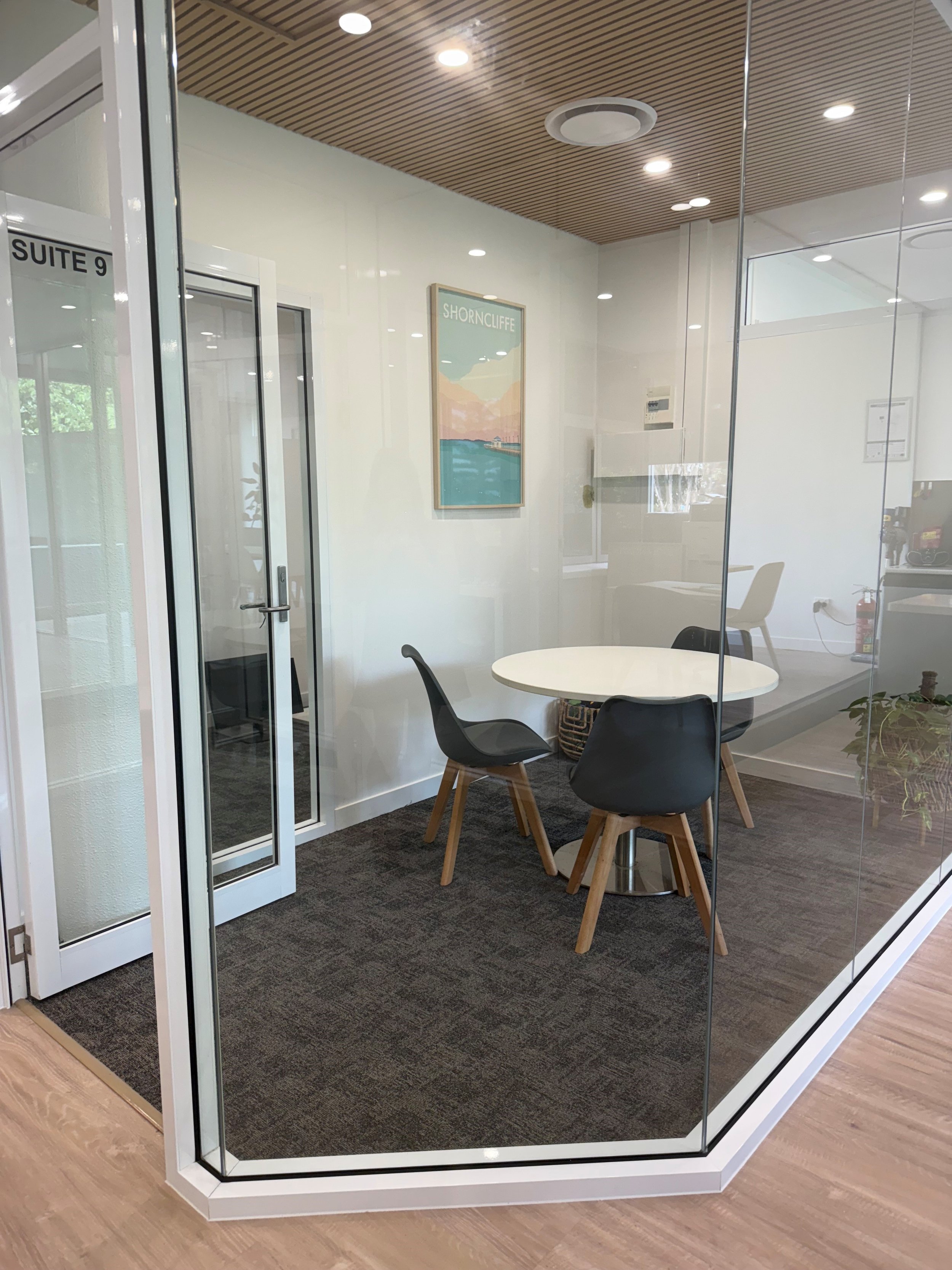 Office conference room with a round table, four black chairs, a white wall with a framed 'Shorncliffe' poster, and glass walls with a door labeled 'Suite 9'.