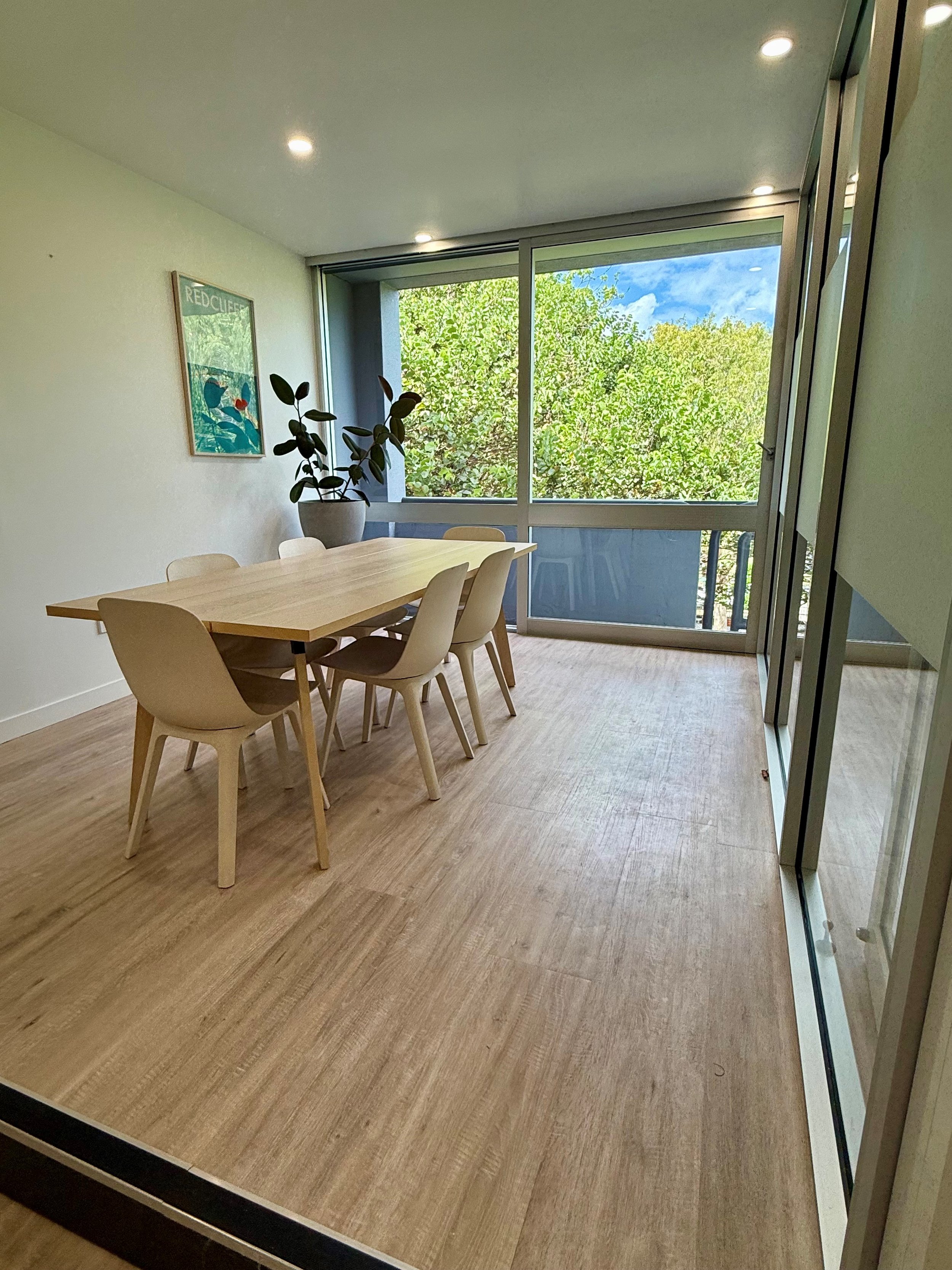 Interior of a modern dining room with a wooden table, six beige chairs, a large potted plant, a framed artwork on the wall, large windows showing green trees outside, and recessed ceiling lights.