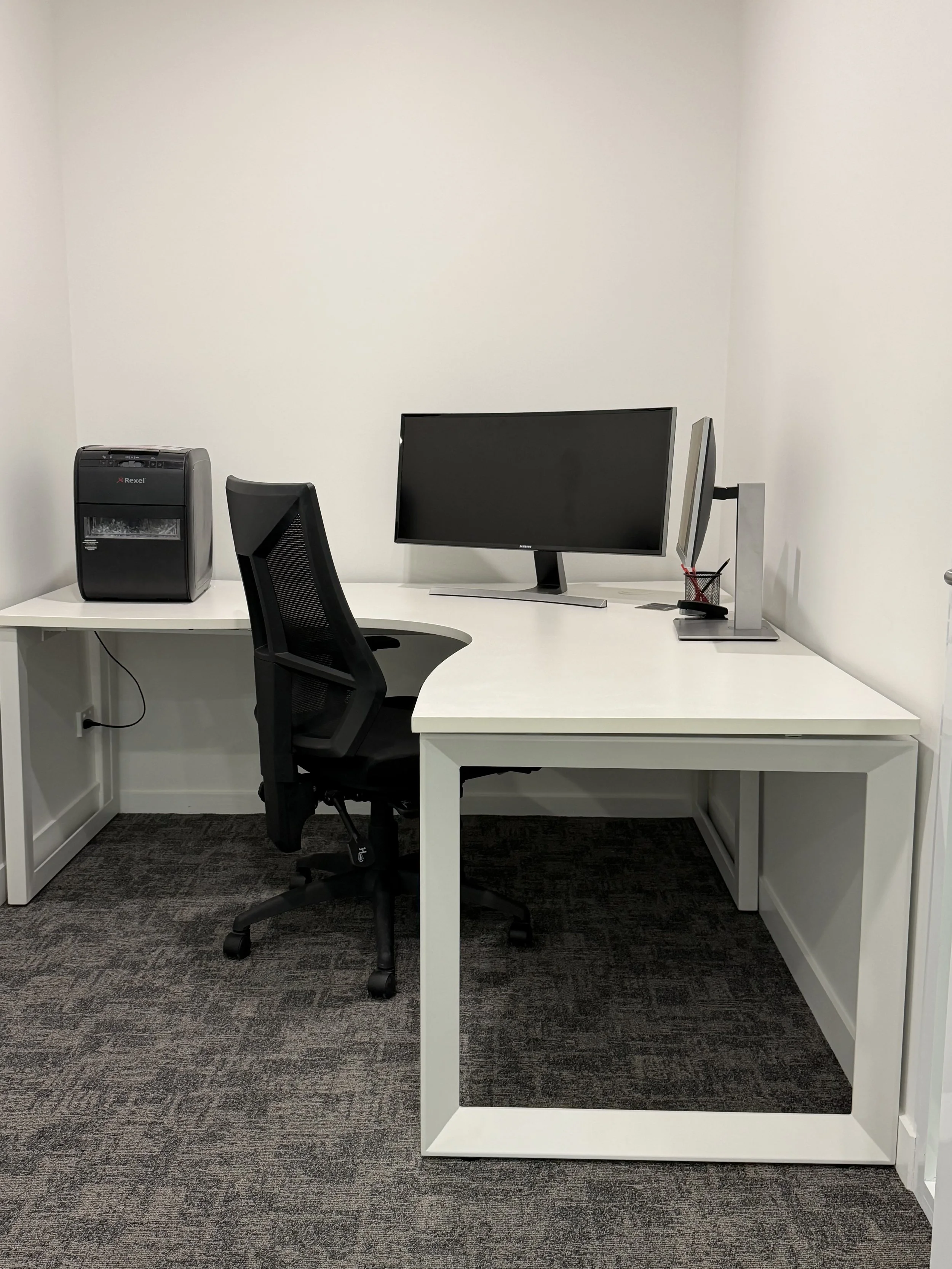 Empty modern office workspace with a white desk, black ergonomic chair, computer monitor, and a shredder on the left, against white walls and gray carpet.