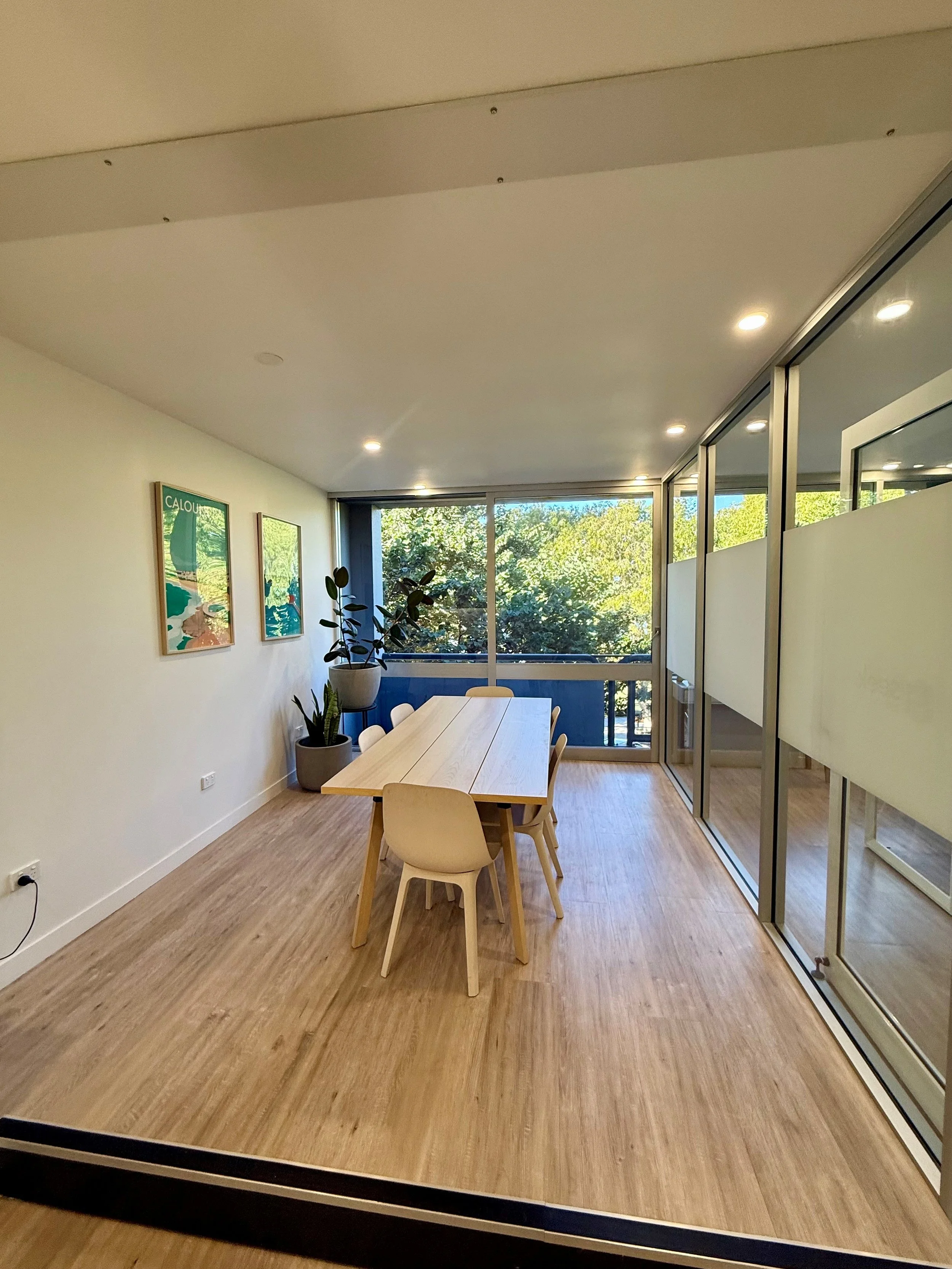An office room with a light wood table and six chairs, several potted plants near a window, framed wall art, and a divider with frosted glass panels. The room has wood flooring and ceiling lights.