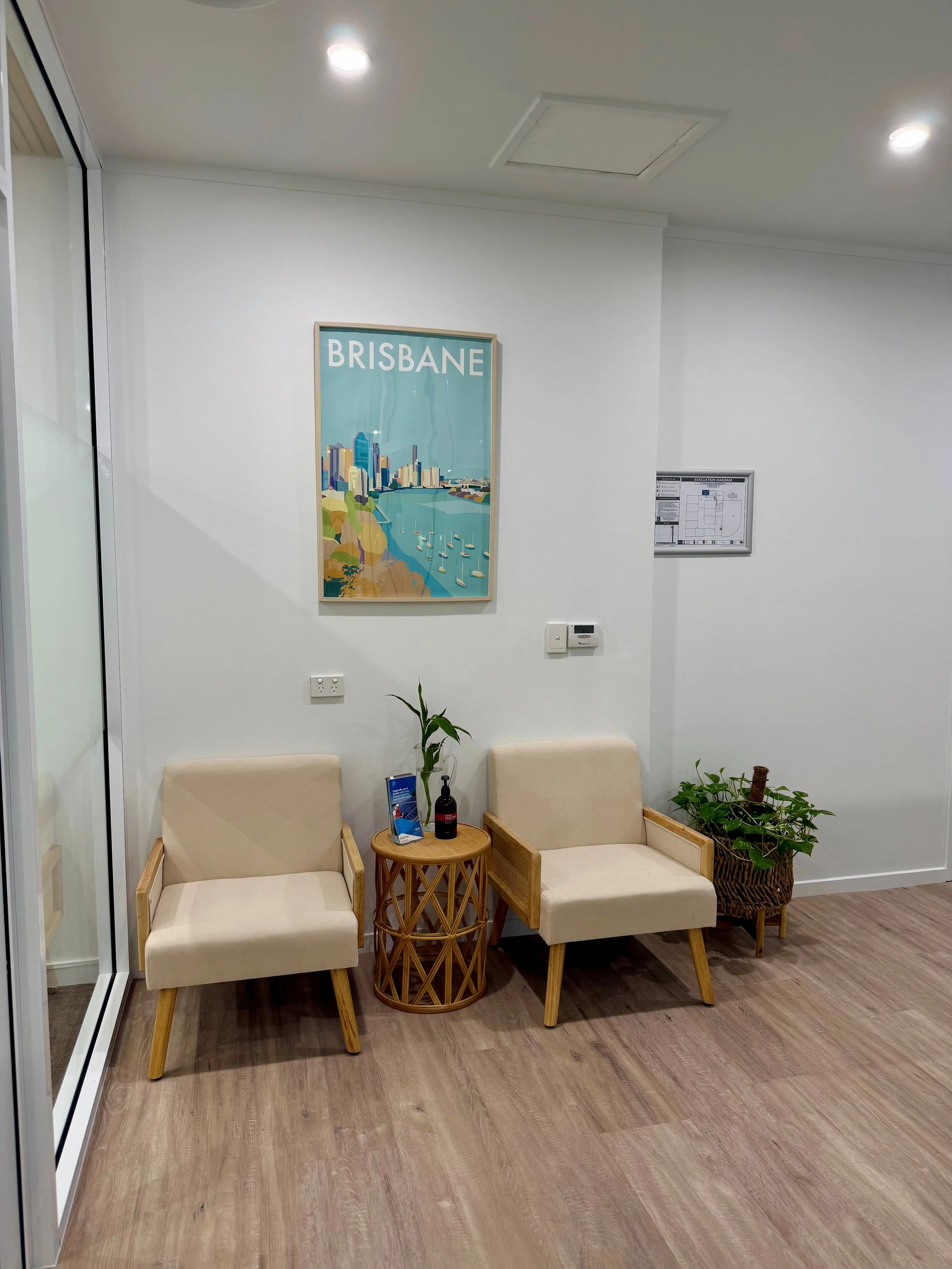 Waiting area with two beige armchairs, a small round wooden side table with bottles and leaflets, a potted plant, and a framed illustration of Brisbane on the white wall.
