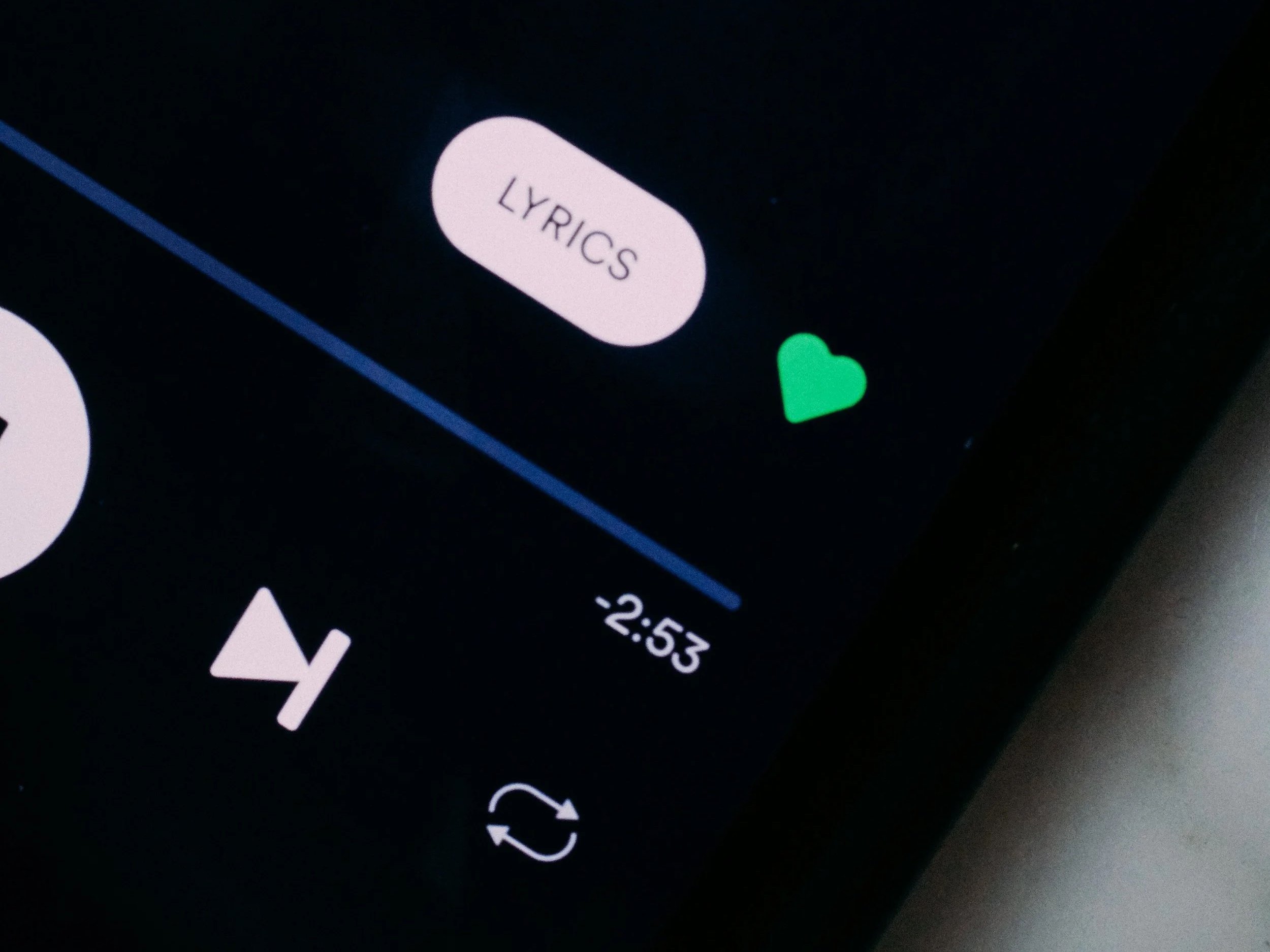 Close-up of a smartphone screen showing the 'LYRICS' button in a music app, with a small green heart icon nearby and part of a white arrow icon at the bottom.