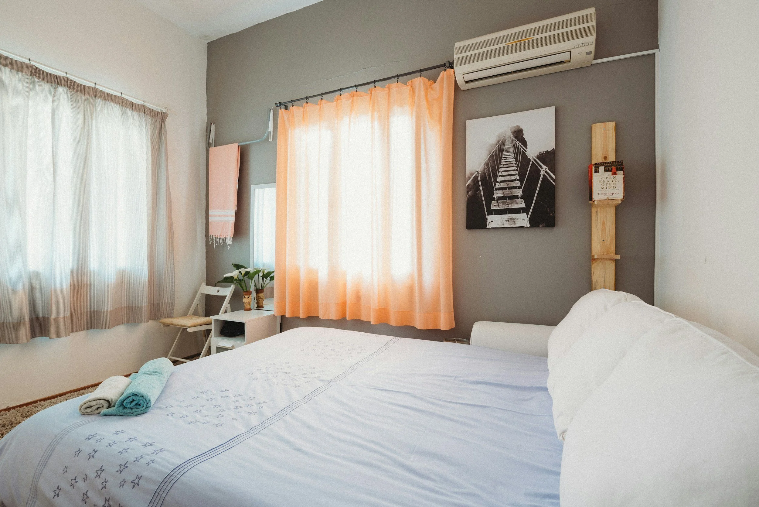 A bedroom with a bed, neatly made, with towels placed on top, and a white side table with plants and decorations. The room has light filtering through sheer curtains and peach-colored curtains, a wall-mounted air conditioner, a black and white photograph of a bridge, and a small wooden bookshelf with books.