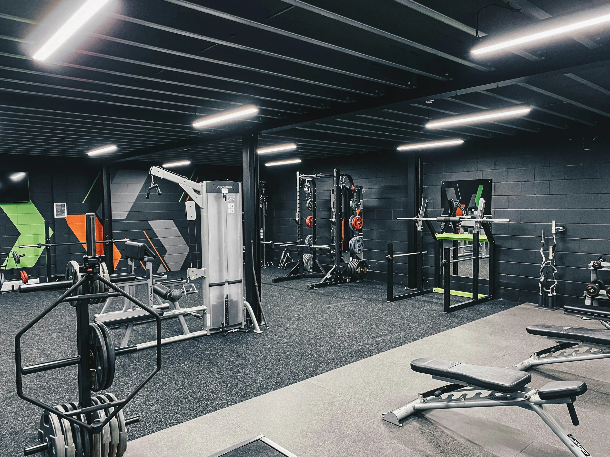 Interior of a modern gym with workout equipment including weight machines, benches, and free weights, with a black and gray color scheme and geometric wall art.
