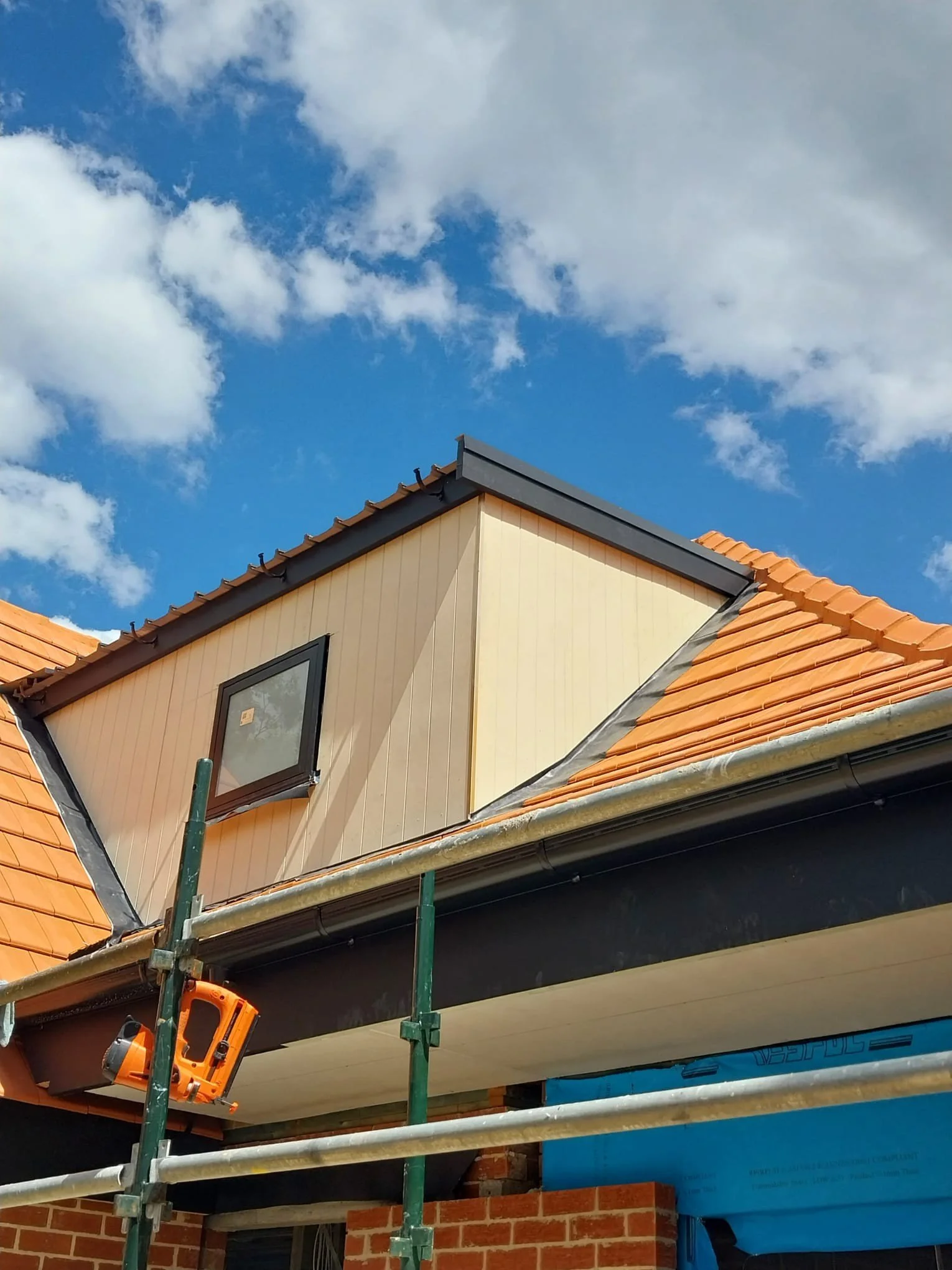 Side view of a house under construction, showing a roof with orange tiles, scaffolding, and a blue sky with some clouds.