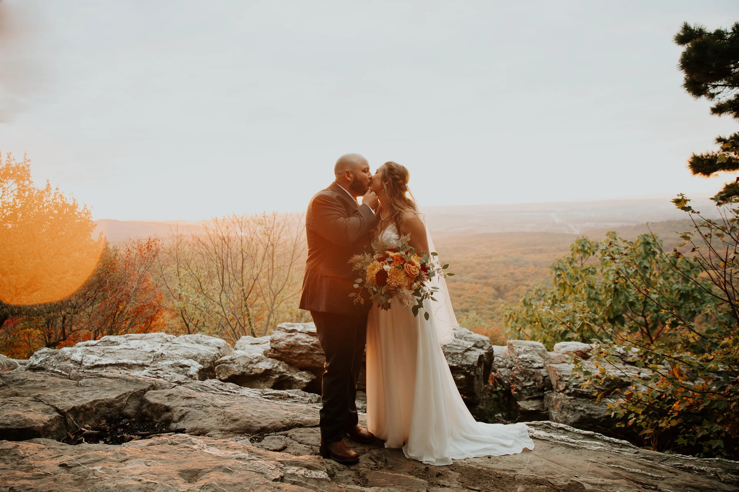An elopement bride and groom share a kiss outdoors on a rocky ledge during sunset, with trees and rolling hills in the background.