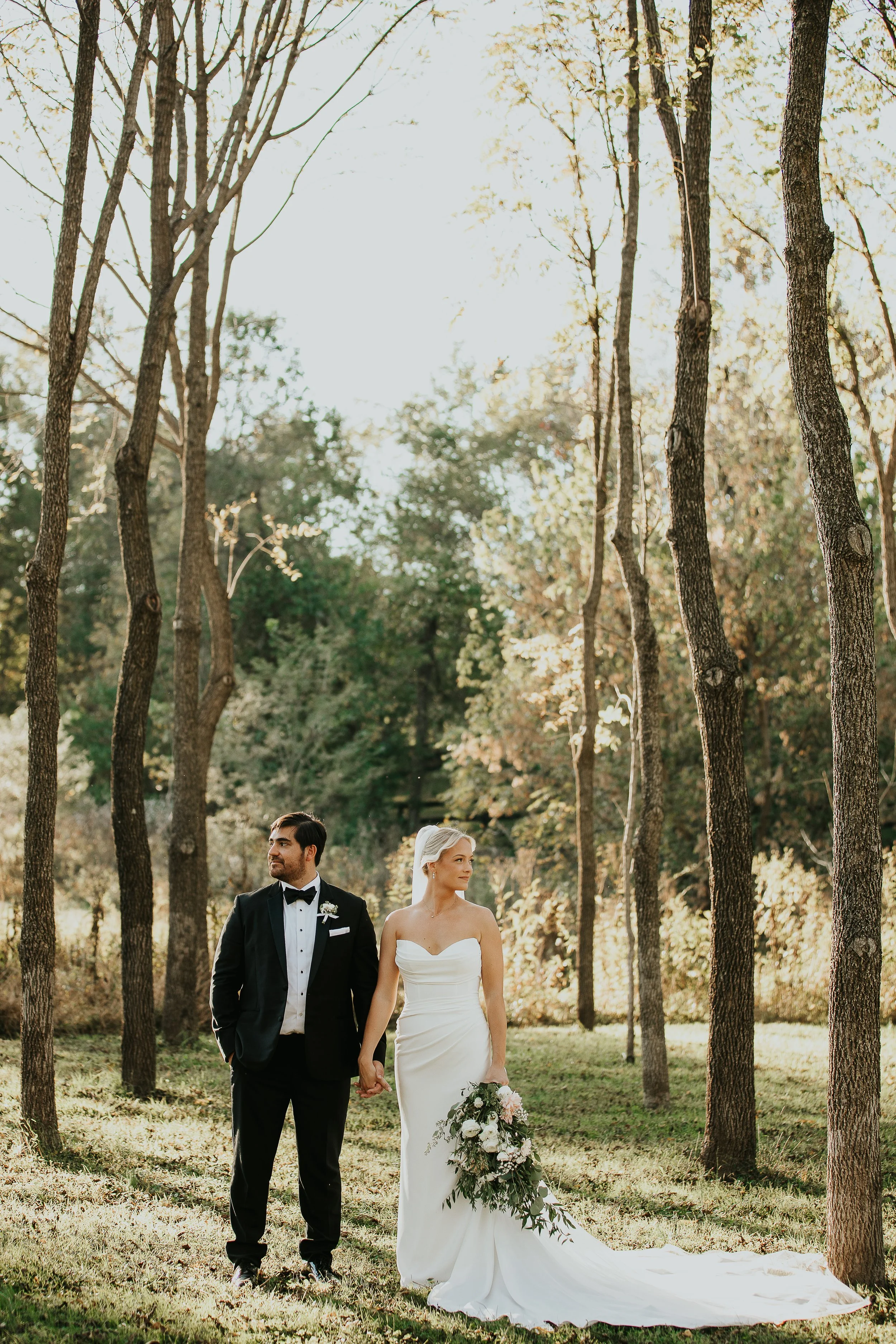 A bride and groom walking hand in hand in a wooded outdoor setting on their wedding day, with sunlight filtering through the trees.
