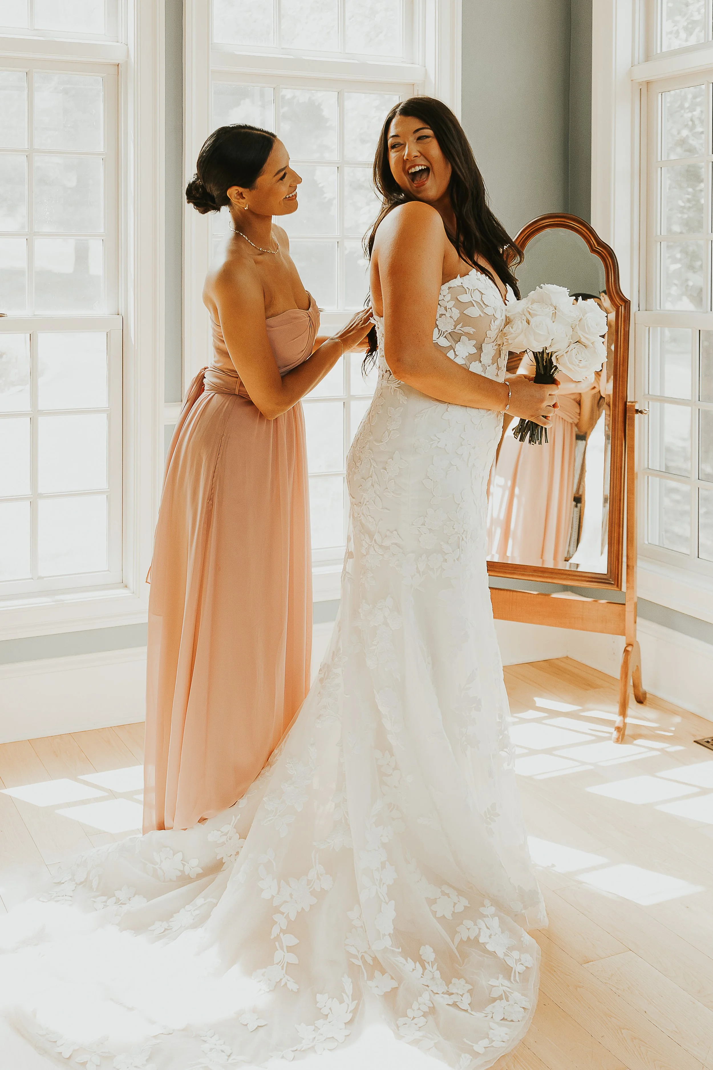 A bride holding a bouquet of white roses, smiling as she looks at her bridesmaid, inside a sunlit room with large windows and a wooden floor.