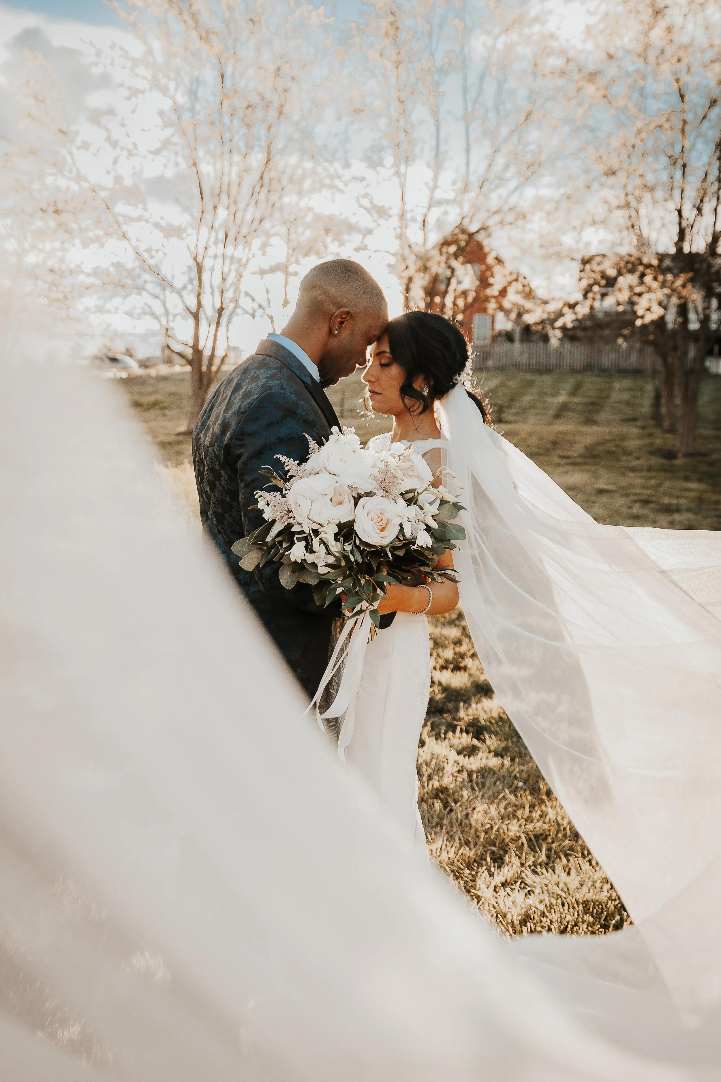 A bride and groom share a tender moment with their foreheads touching, outdoors during sunset. The bride holds a bouquet of white roses and greenery. The background features blooming trees and a fence, with warm, golden lighting.
