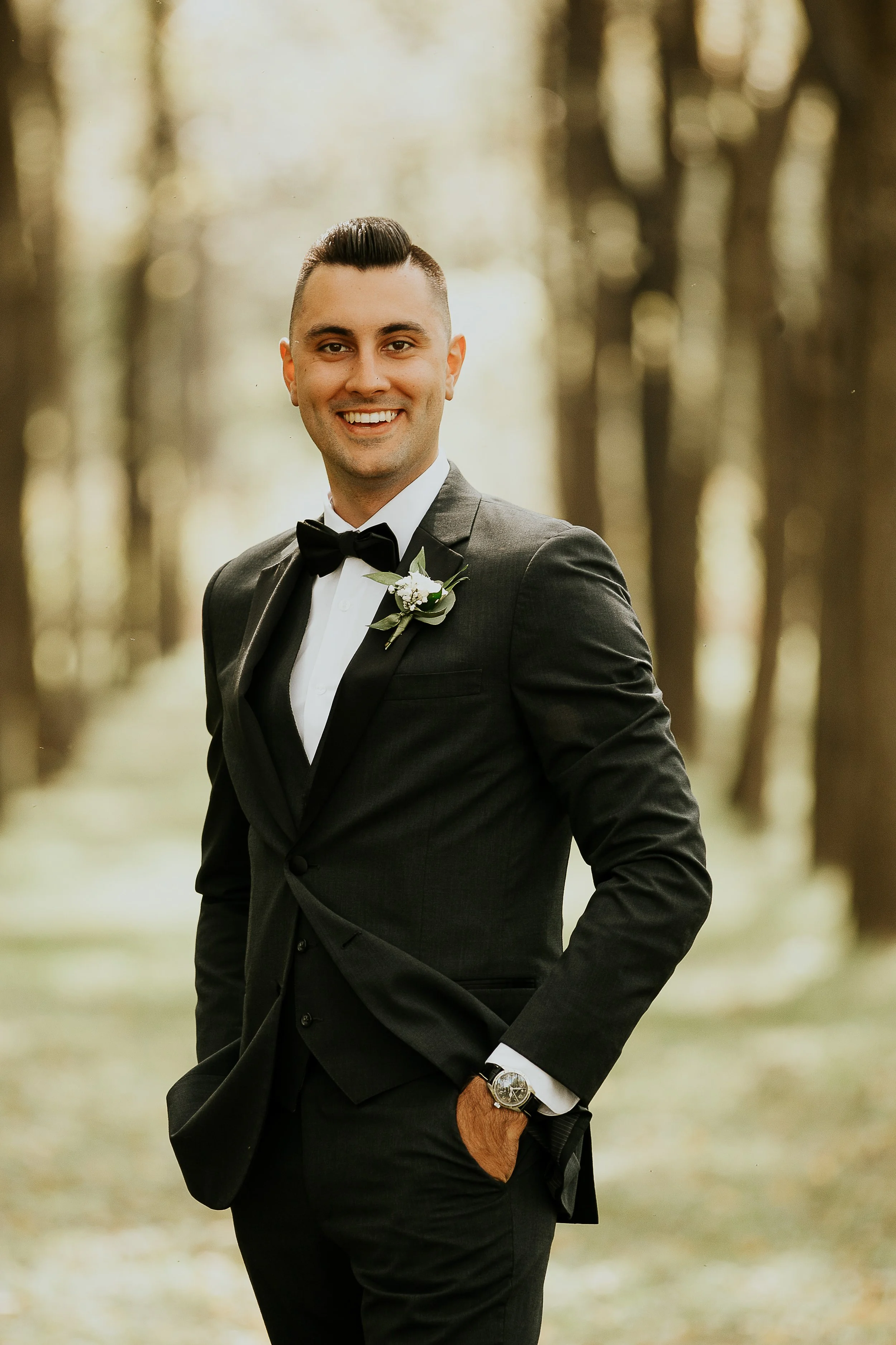 A man in a tuxedo with a boutonniere, smiling outdoors in a wooded area with blurred trees in the background.