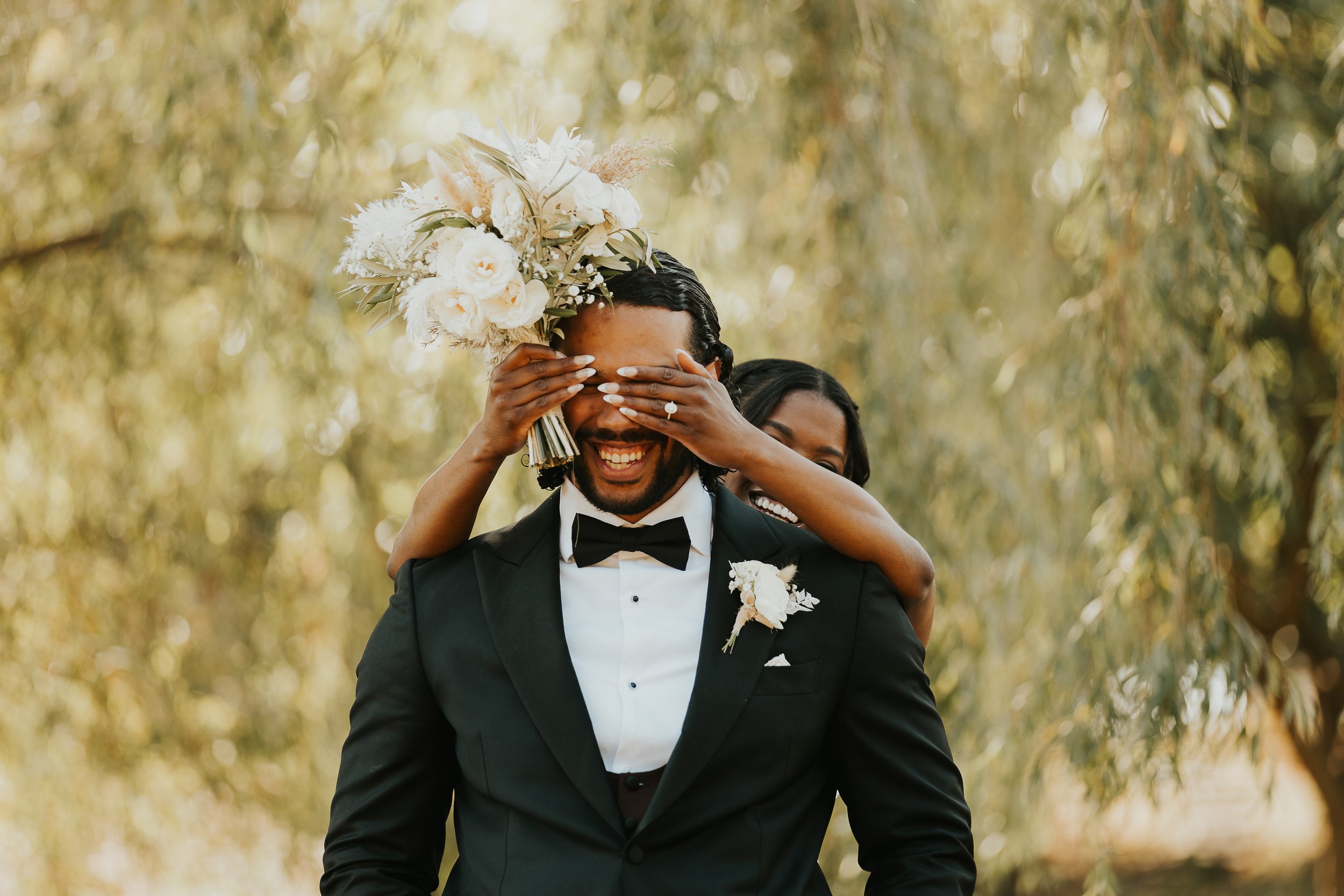 A joyful man in a black tuxedo with a bow tie, carrying a bouquet of white flowers on his head, while a woman smiling behind him playfully covers his eyes with her hands. They are outdoors with a blurred background of trees and sunlight.