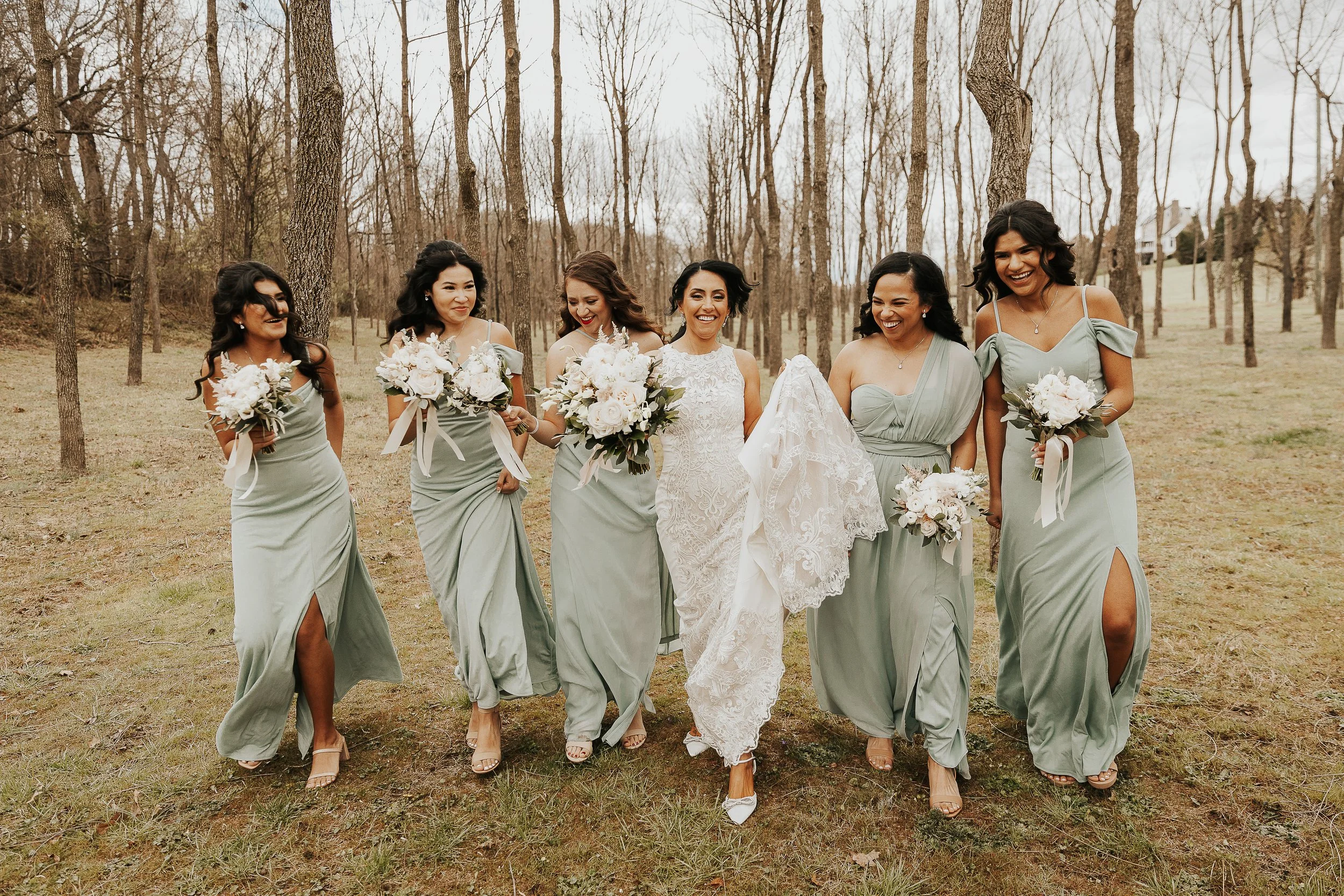 A bride and six bridesmaids walking together outdoors on a grassy area with leafless trees in the background. The women are smiling and holding bouquets of flowers.