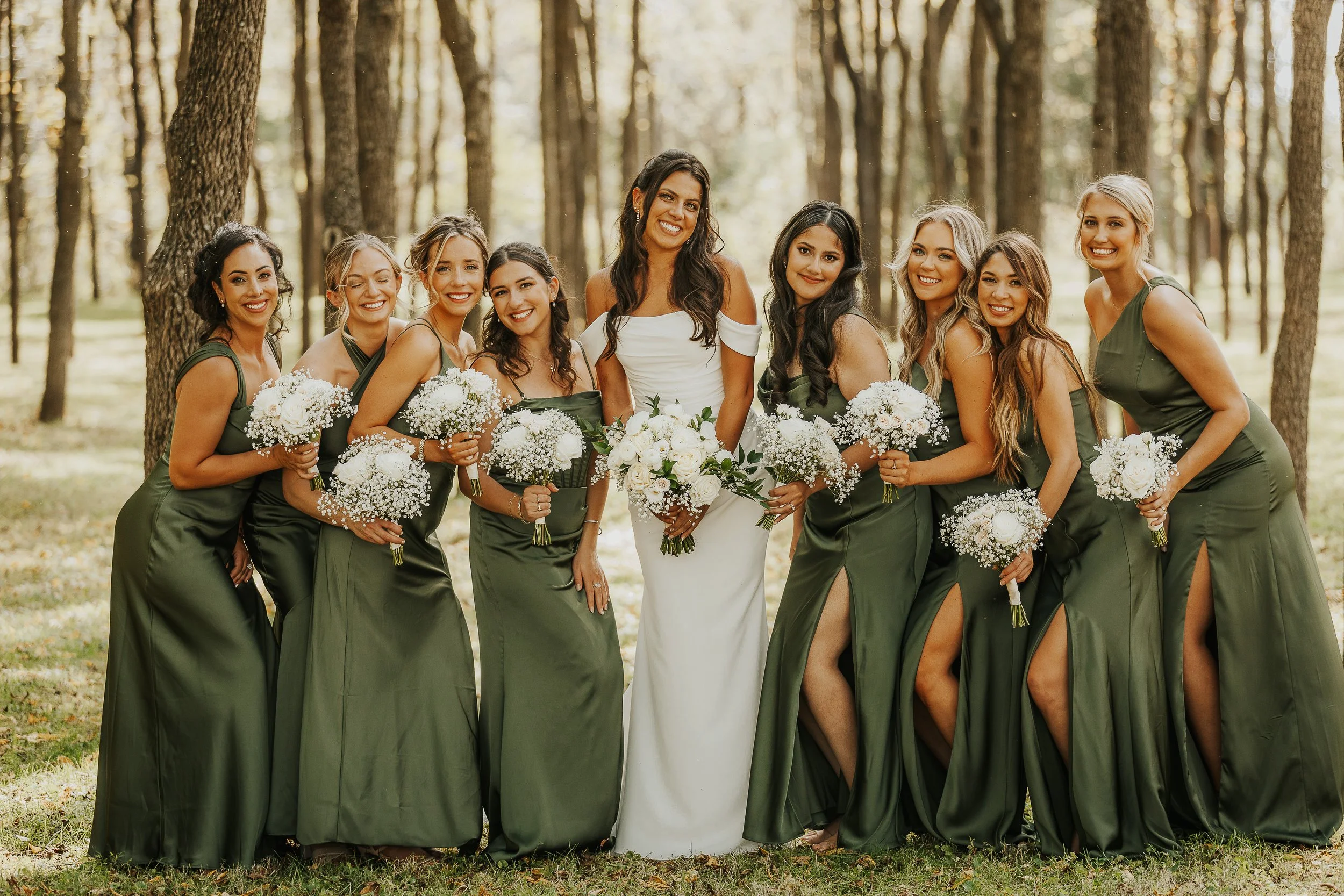 A group of women dressed in green bridesmaid dresses and a bride in a white gown, standing outdoors among tall trees, holding bouquets of white flowers, smiling at the camera.