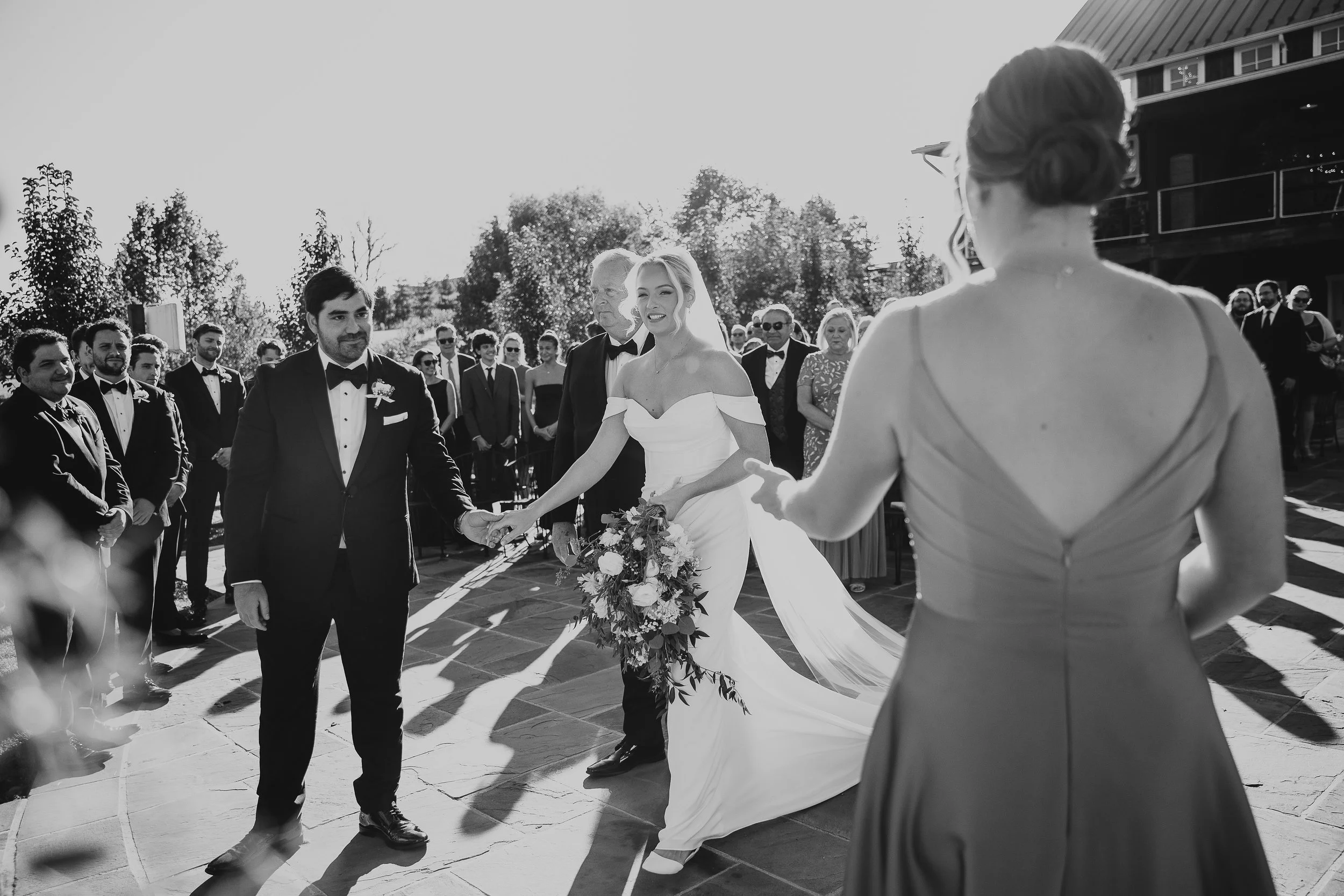 Black and white photo of a wedding ceremony outdoors, with the bride holding a bouquet and the groom holding her hand, surrounded by wedding guests.