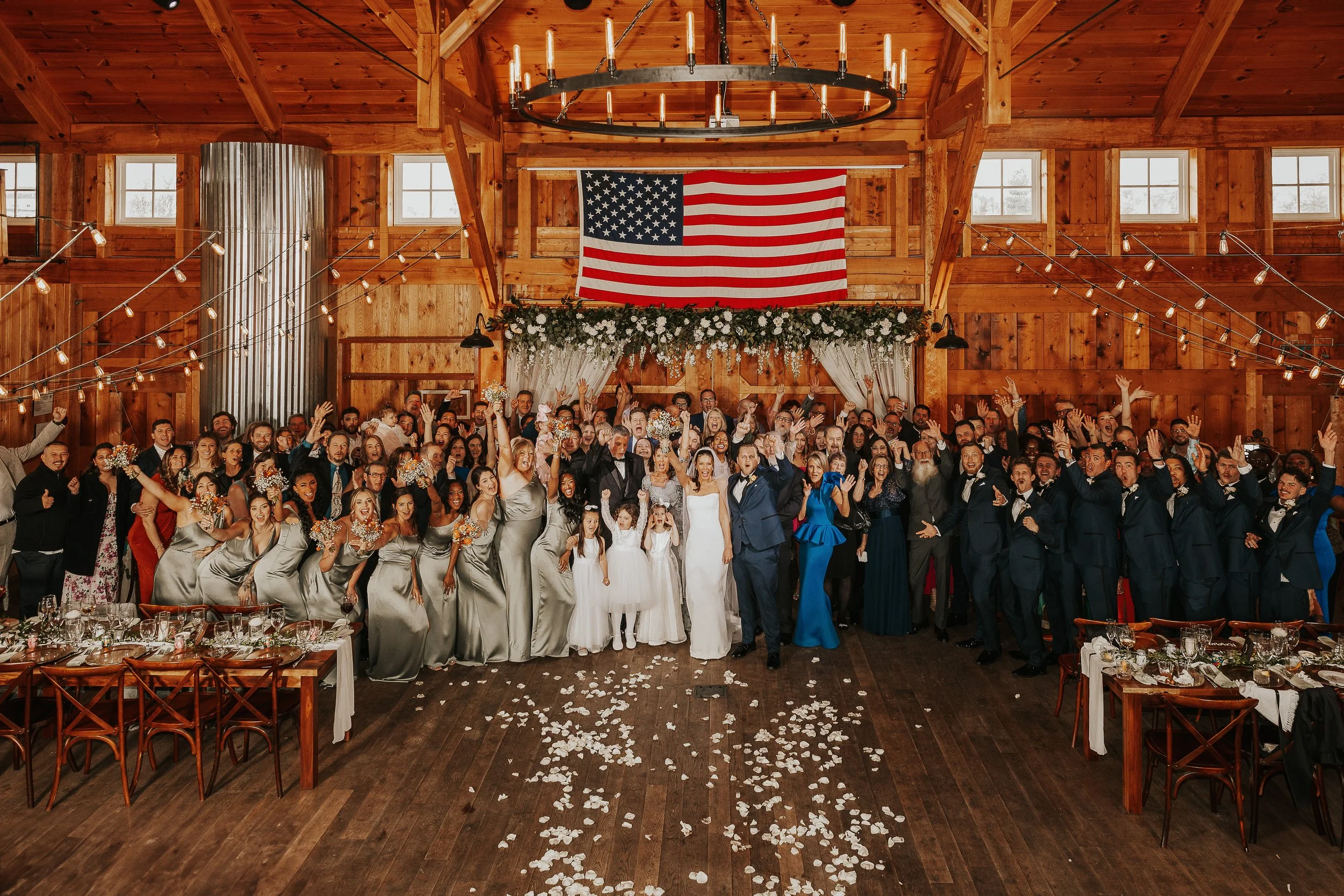 A large group of wedding guests celebrating inside a rustic wooden venue with string lights, American flag, and floral decorations, with flower petals on the floor.