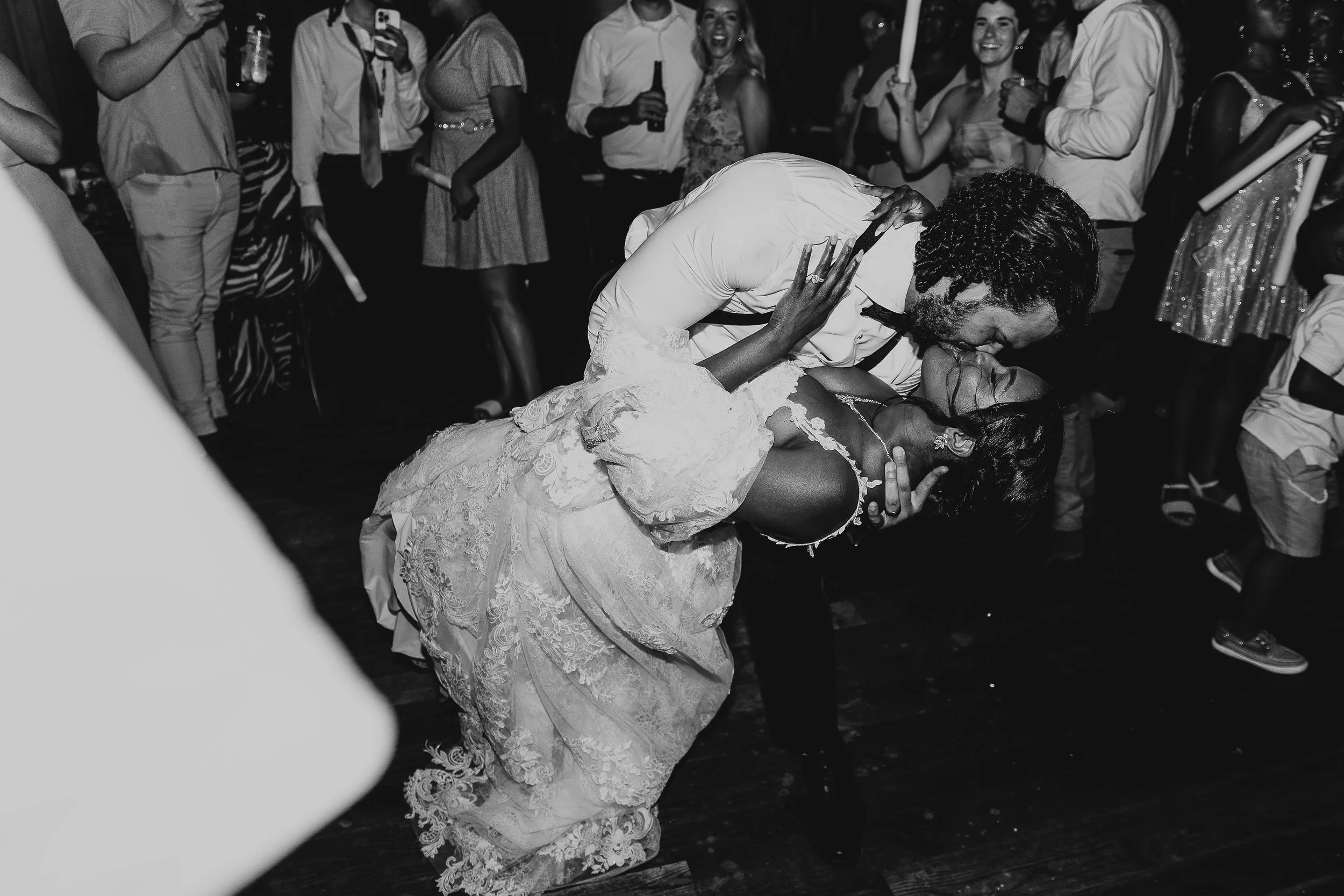 Black and white photo of a wedding reception with a couple dancing, the bride wearing a lace wedding dress and the groom wearing a suit. The groom is dipping the bride while kissing her, and they are surrounded by guests who are watching, laughing, a