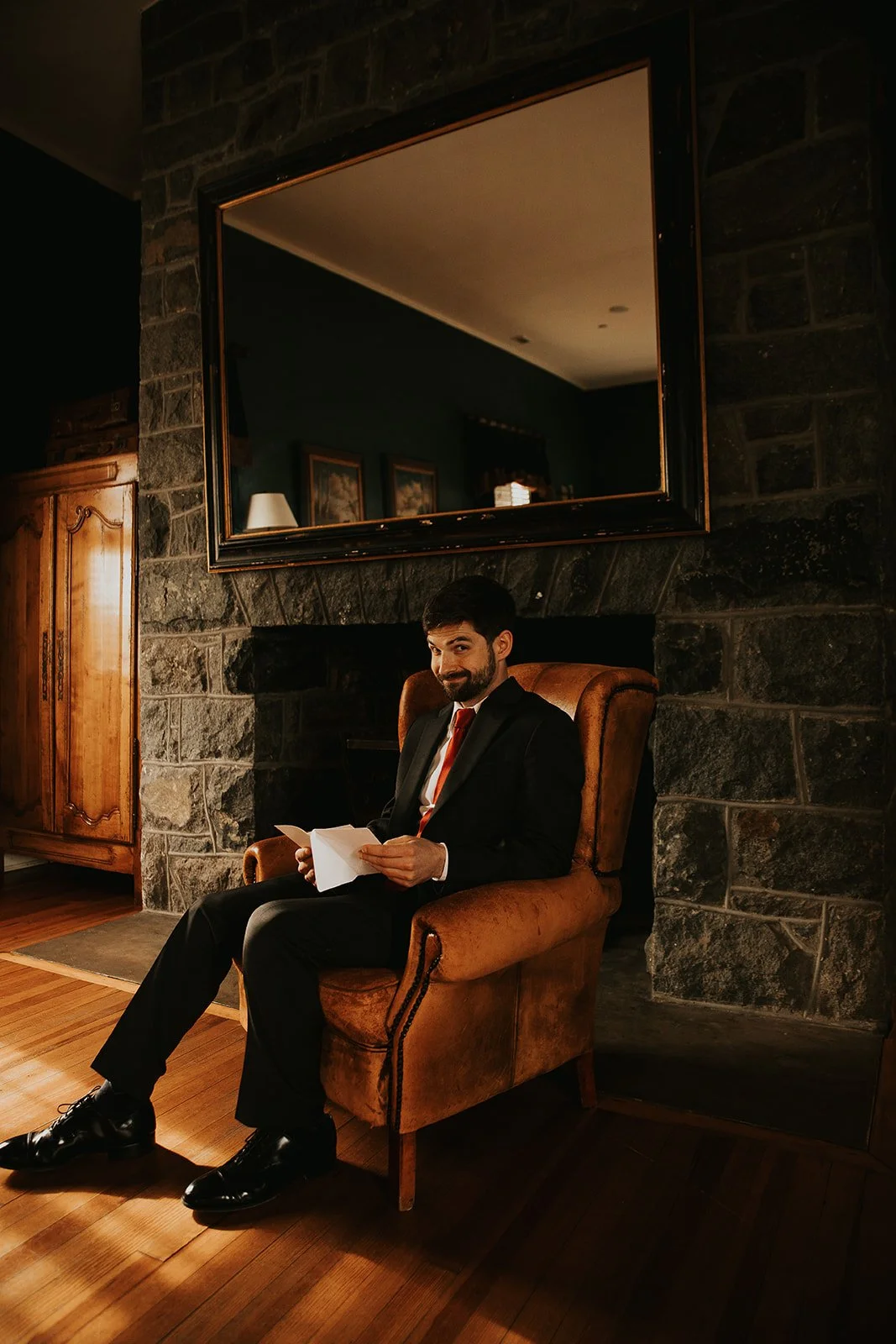 A man in a suit sitting in a brown armchair, reading a letter, in a room with stone walls, a large mirror, and wooden furniture.