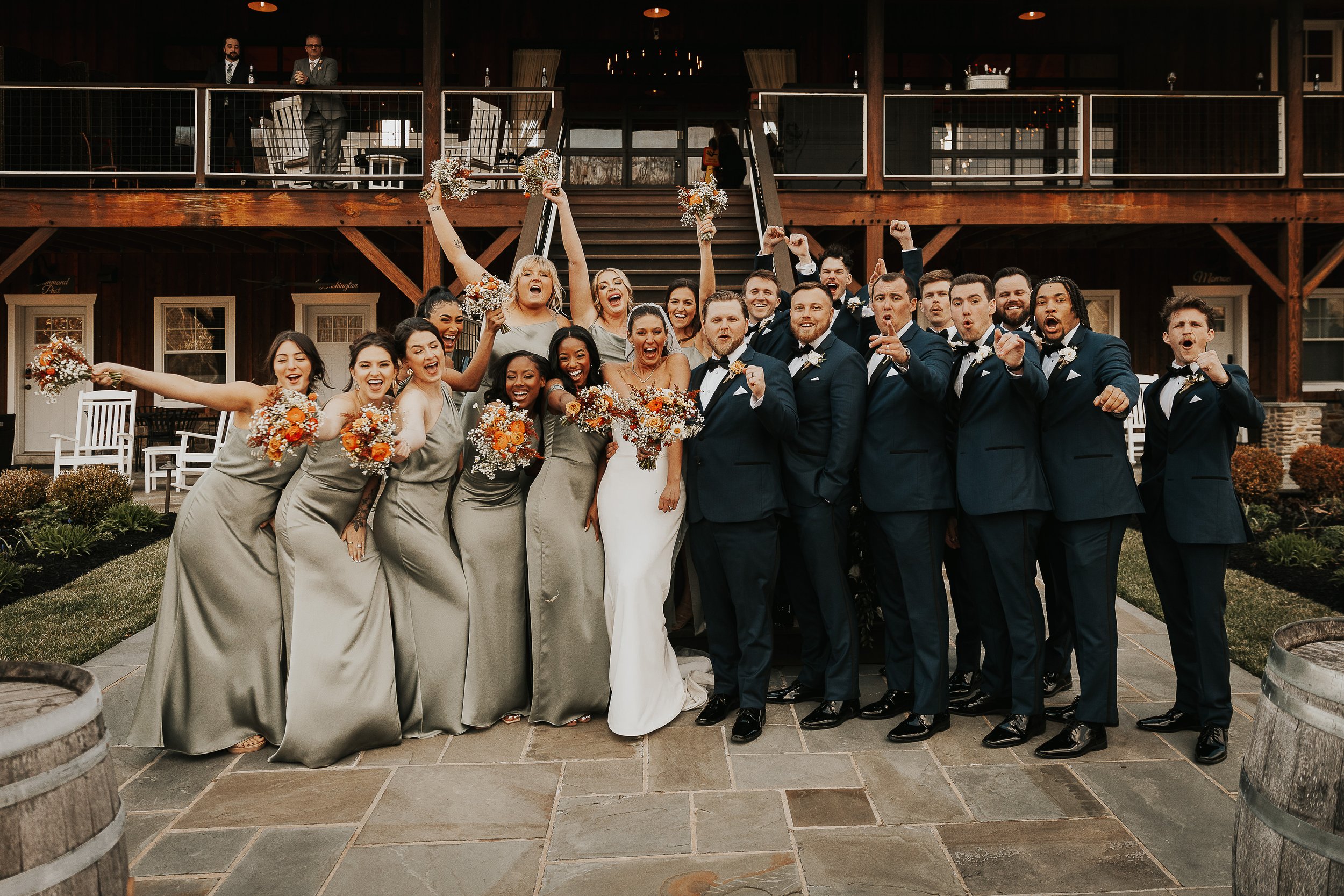 Wedding party celebrating outdoors, with women in matching champagne dresses holding bouquets and men in tuxedos, some raising fists and expressing excitement in front of a rustic building.