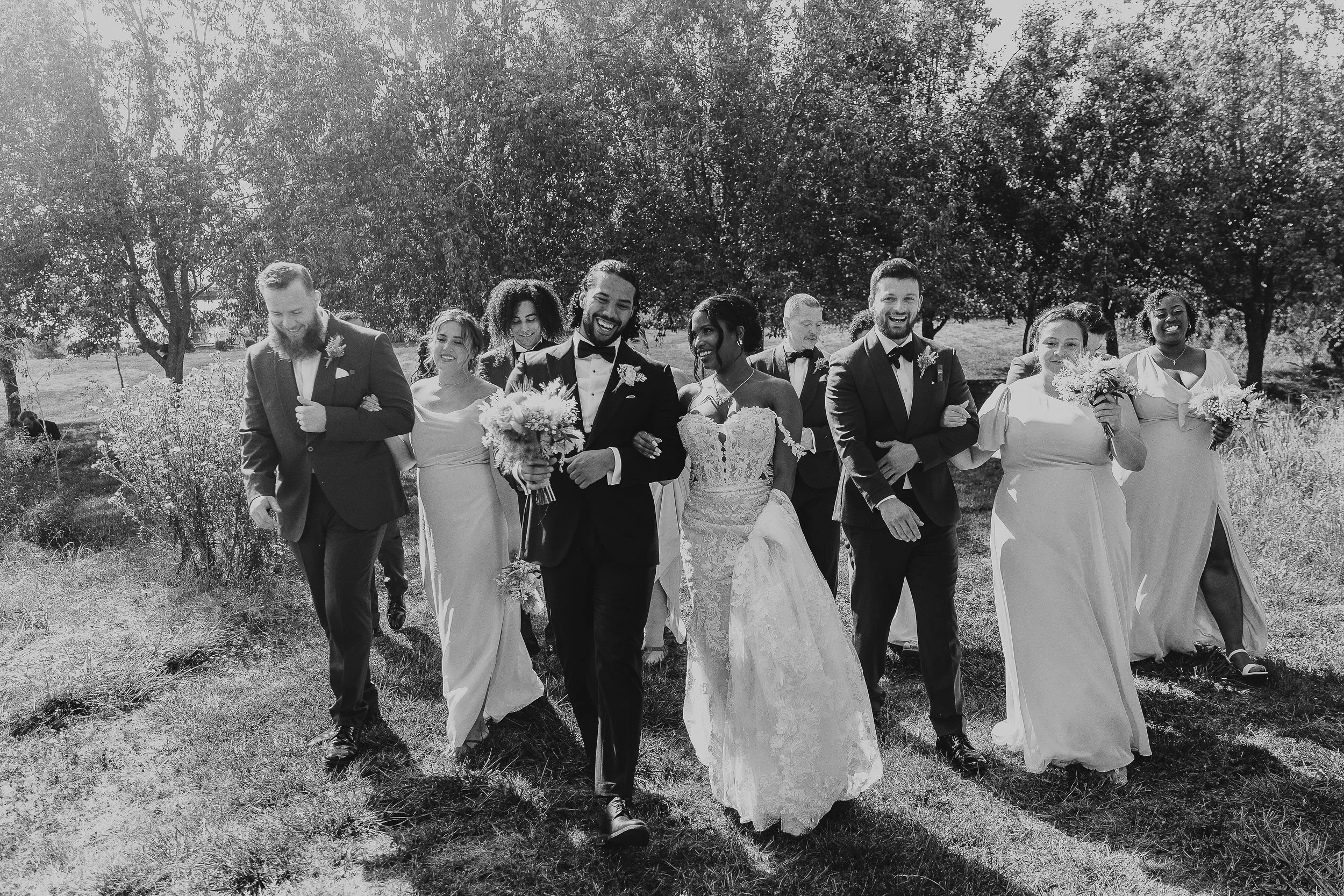 A group of diverse people dressed in formal attire, walking outdoors in a grassy area with trees, smiling and holding bouquets, likely at a wedding celebration.