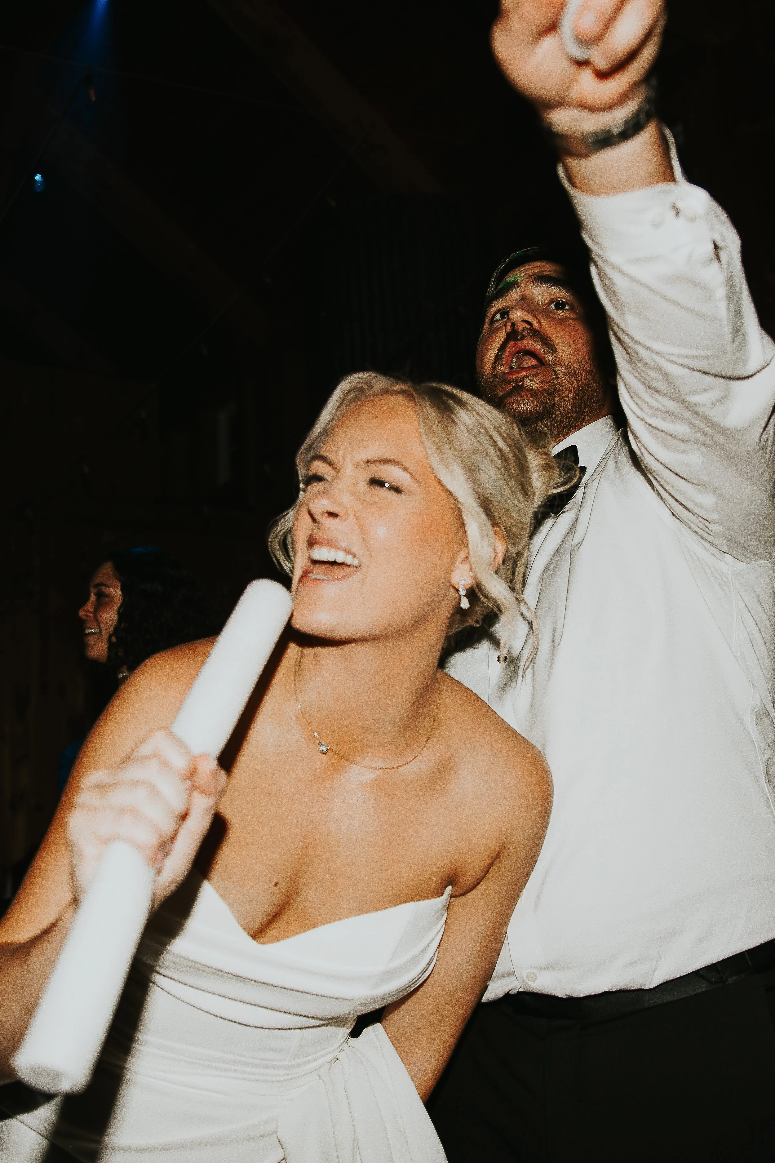 A woman in a white dress holding a microphone, smiling and dancing at a celebration or party with a man in a white shirt.