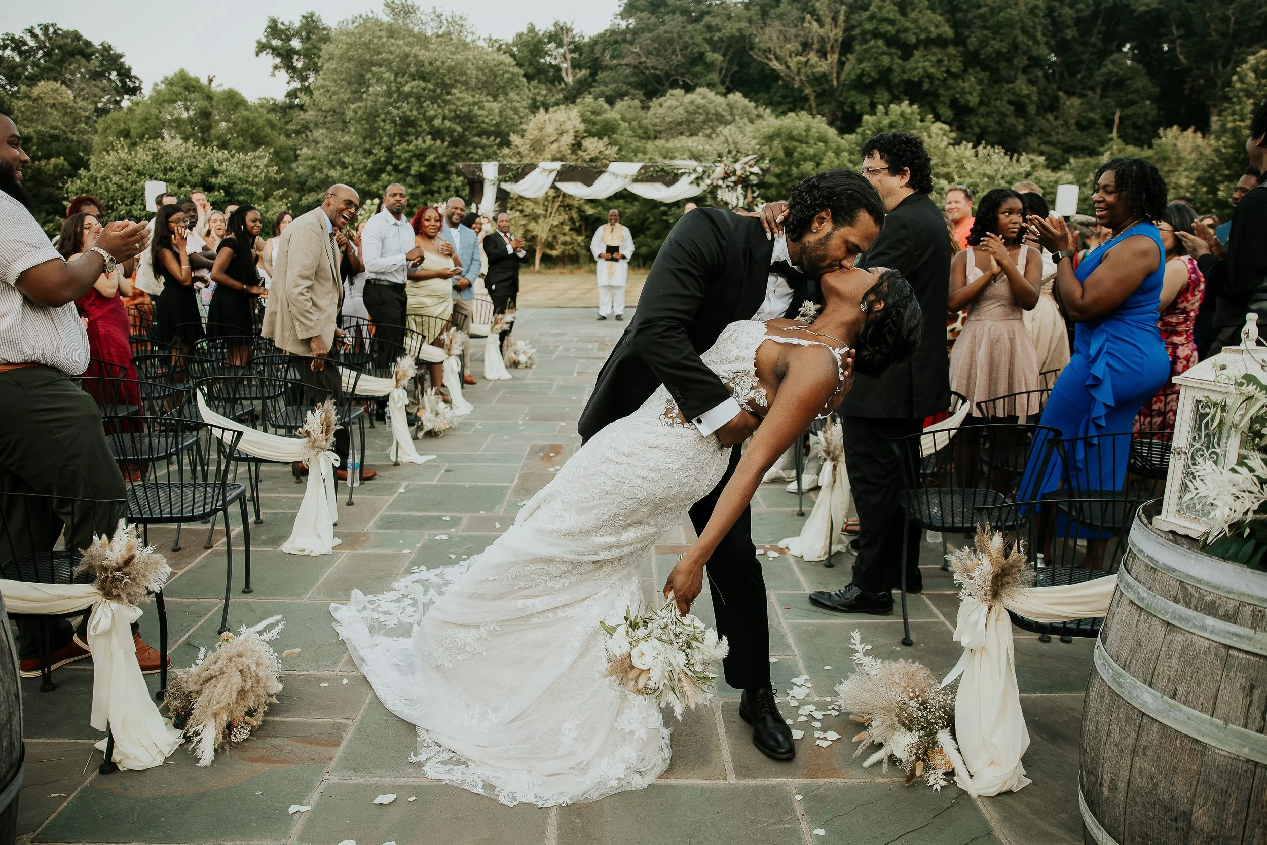 A newly married couple is sharing a romantic kiss during their outdoor wedding ceremony, surrounded by guests and decorative floral arrangements.