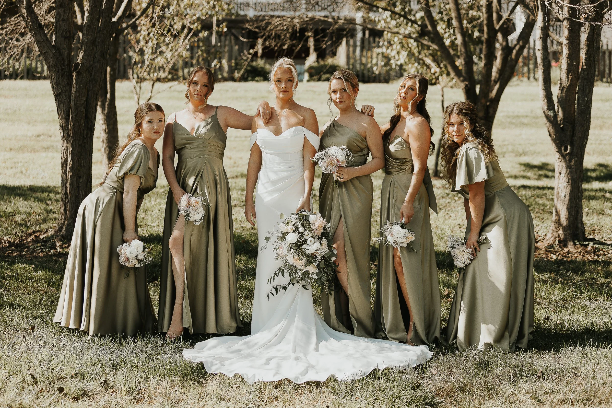 Bride and six bridesmaids standing outdoors under trees during a wedding photoshoot. The bride wears a white strapless wedding gown, holding a bouquet, and the bridesmaids wear matching olive green dresses, each holding a small bouquet.