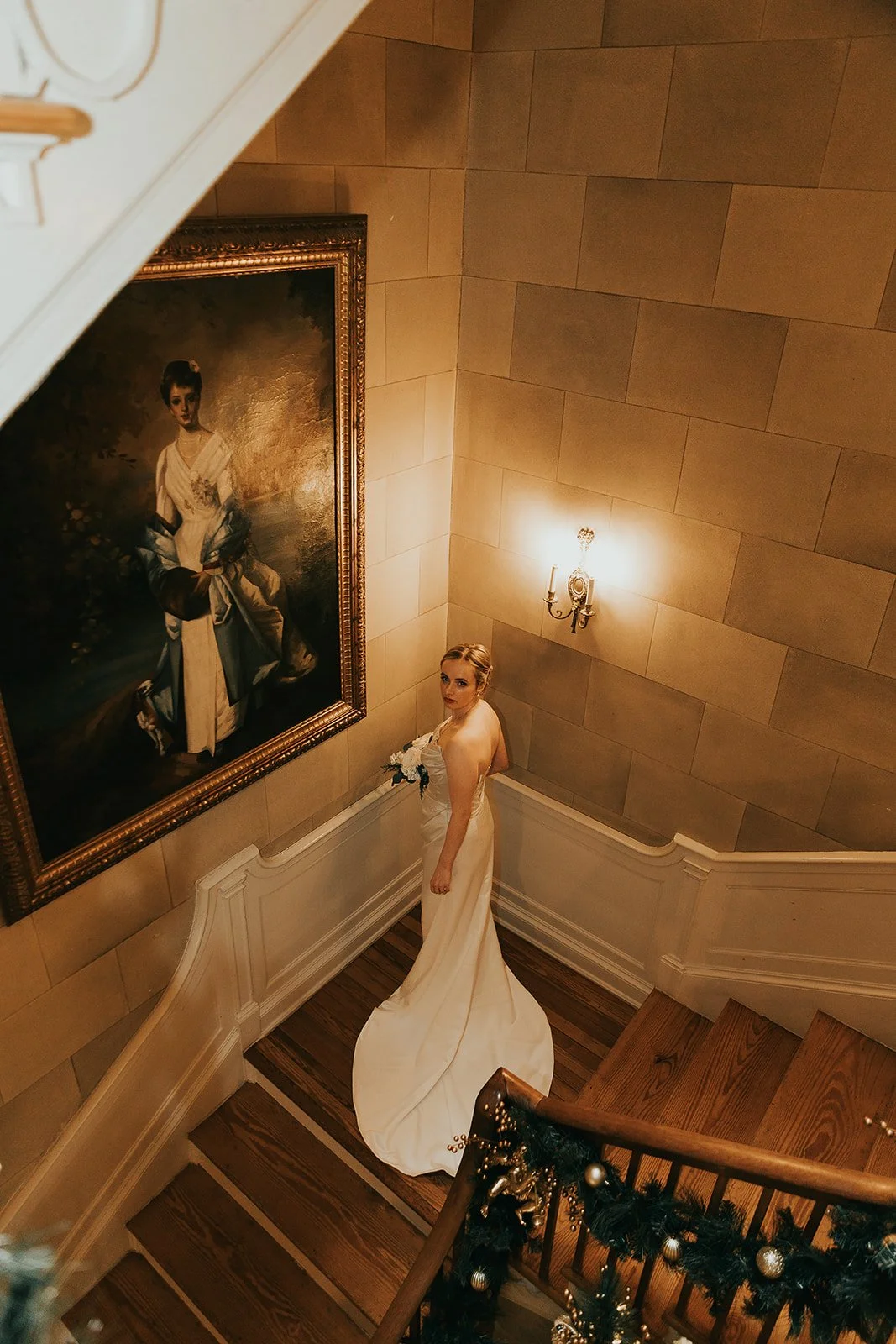 A woman in a white wedding dress holding a bouquet, standing on a wooden staircase in a decorated interior. A large portrait painting and a wall sconce light are visible on the beige tiled wall.