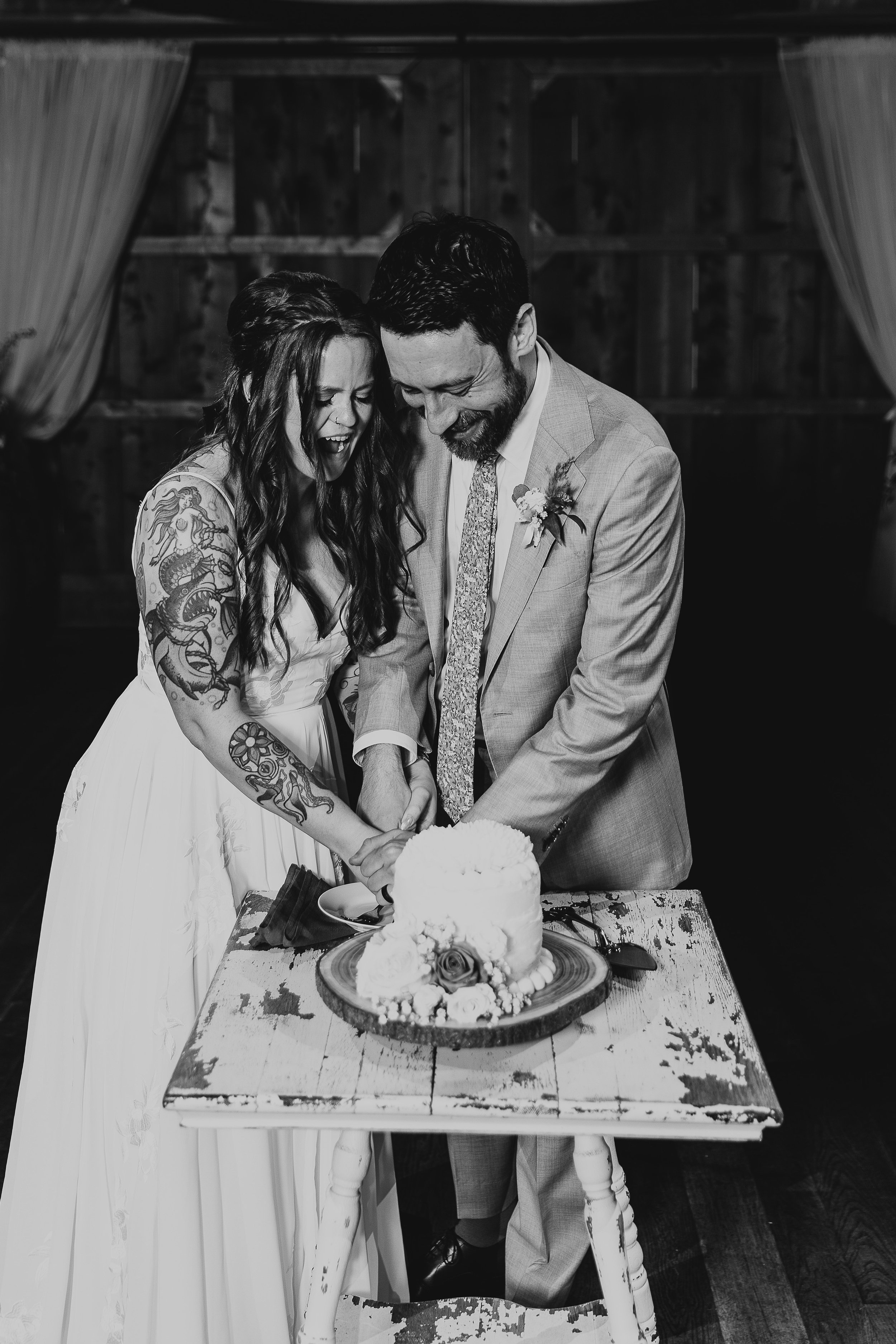 A bride and groom smiling and laughing as they cut a wedding cake together, with a rustic wooden background and table.