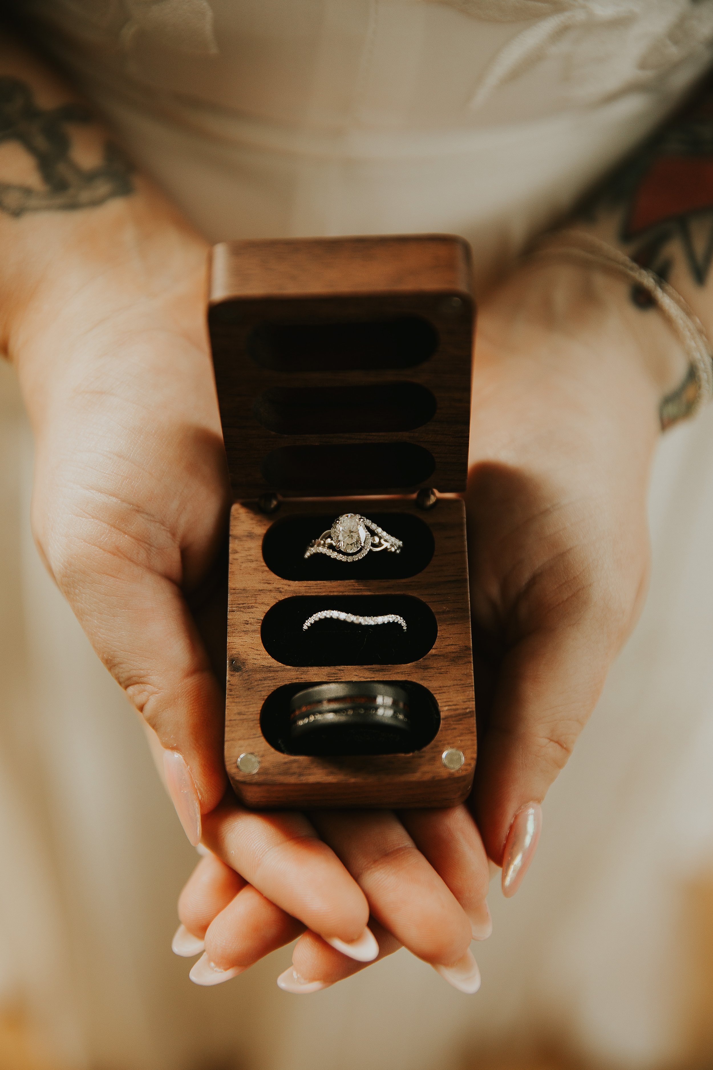 A person holding an open small wooden ring box with a diamond engagement ring inside. The person has manicured nails and visible tattoos on their arms.